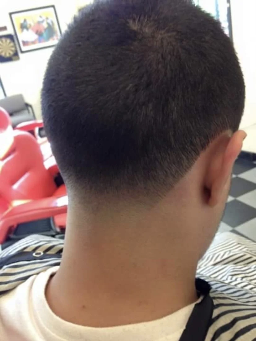 Back of a person's head with a fresh fade haircut, short dark hair, and a striped shirt, sitting in a barbershop or salon with red chairs and posters on the wall.