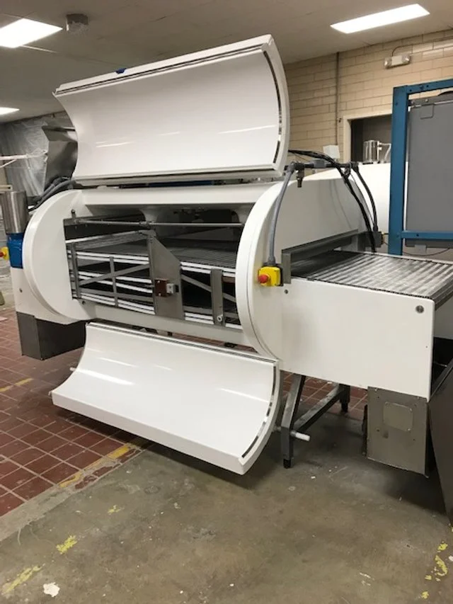 Large industrial machine with a white curved cover, metal rollers, and a conveyor belt in a factory setting.