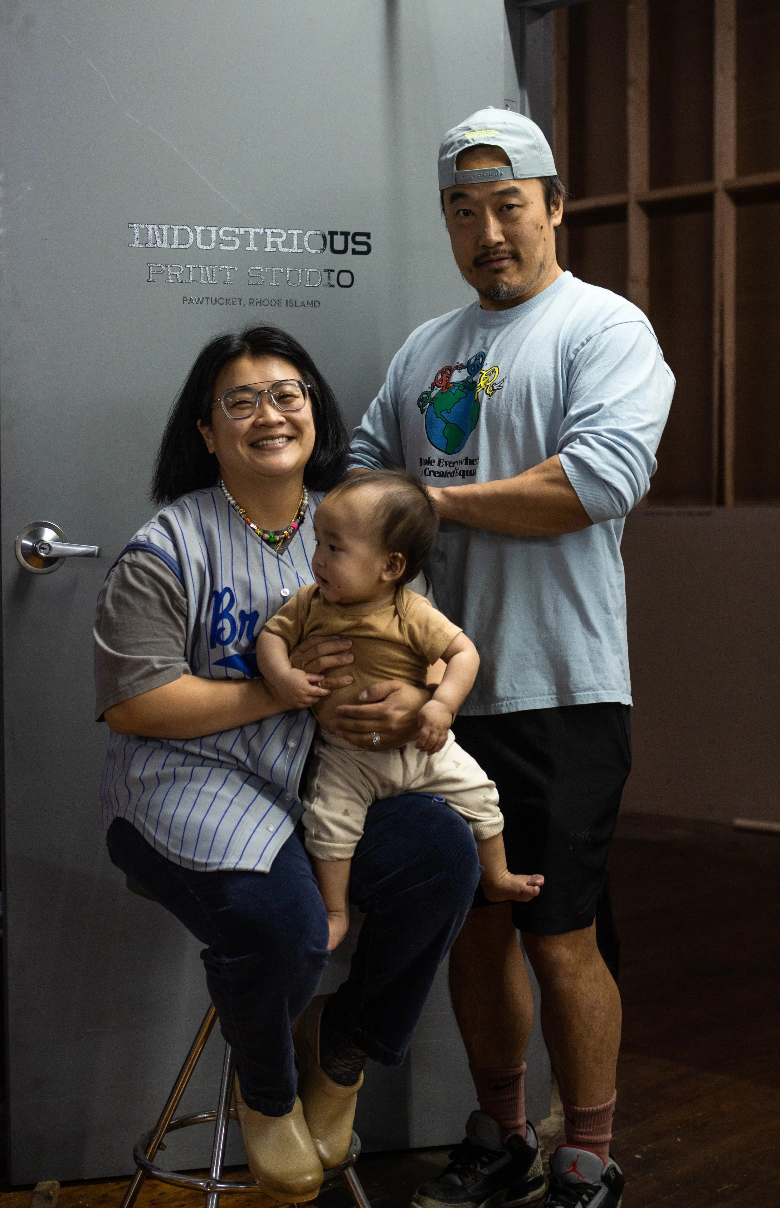 Three people, two adults (Brad and Liz Czarnowski, owners)  and a baby, in front of a gray door with 'INDUSTRIOUS STUDIO' written on it. The woman is holding the baby, and the man is standing next to them.