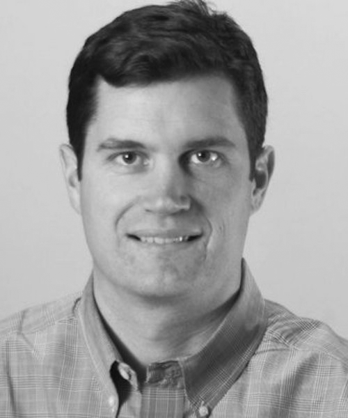 Black and white portrait of a man with short dark hair, wearing a button-down shirt, smiling slightly.
