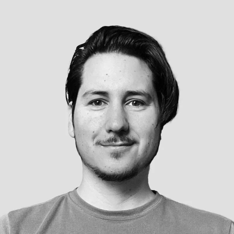 Black and white portrait of a smiling young man with short, slicked-back hair, facial hair, and wearing a casual t-shirt against a plain background.
