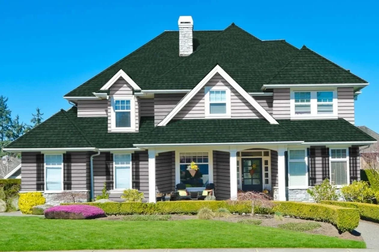 Front view of a large two-story house with purple siding, white trim, multiple windows, a porch with white columns, a green shingle roof, and a well-maintained lawn with bushes and flowers.