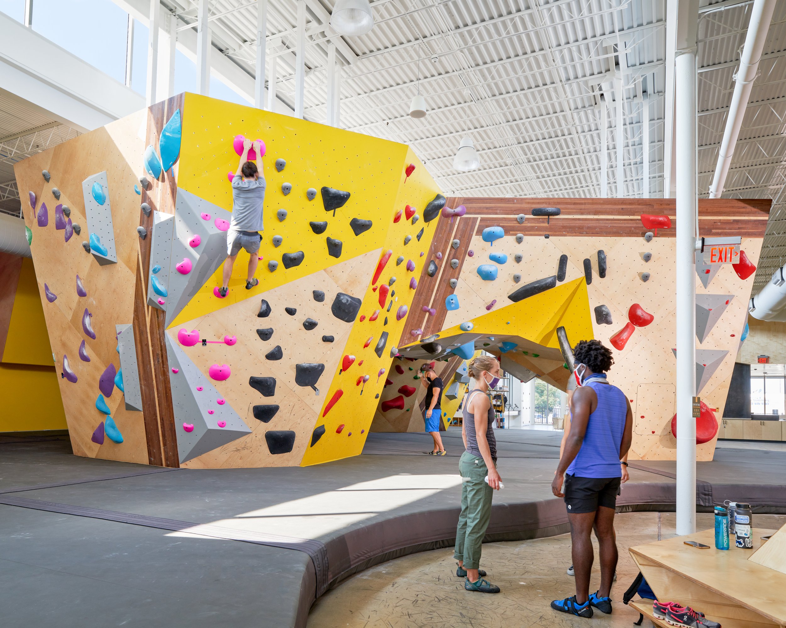 This view captures the dynamic geometry of Crux Central’s climbing walls, where angled plywood surfaces and vibrant holds support movement, conversation, and community in a sunlit setting.