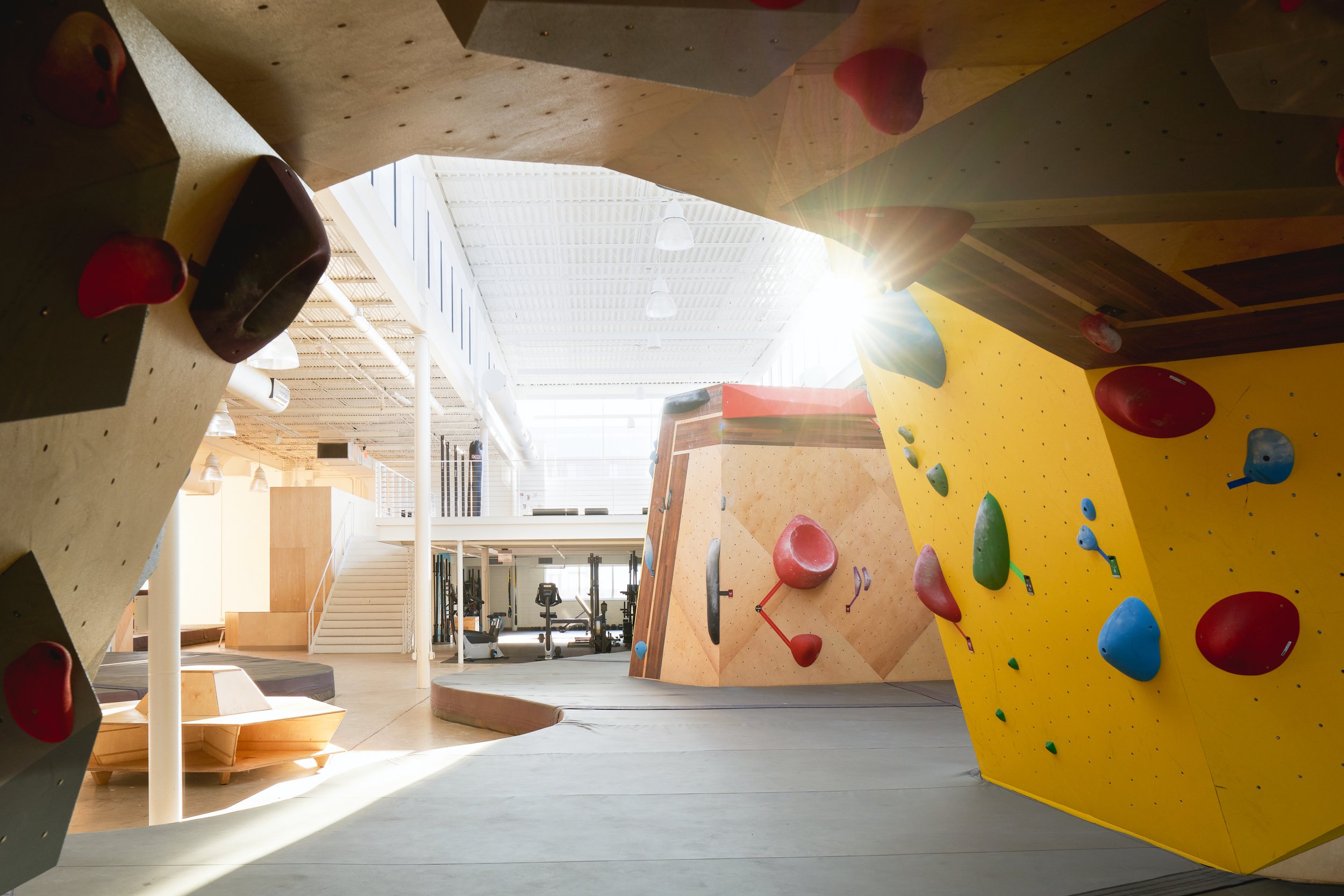 Captured from a tucked-in angle, this shot frames the morning sun bursting through the clerestory—casting soft light across bouldering walls, stairs, and gym equipment. Architecture and activity meet in layered depth.