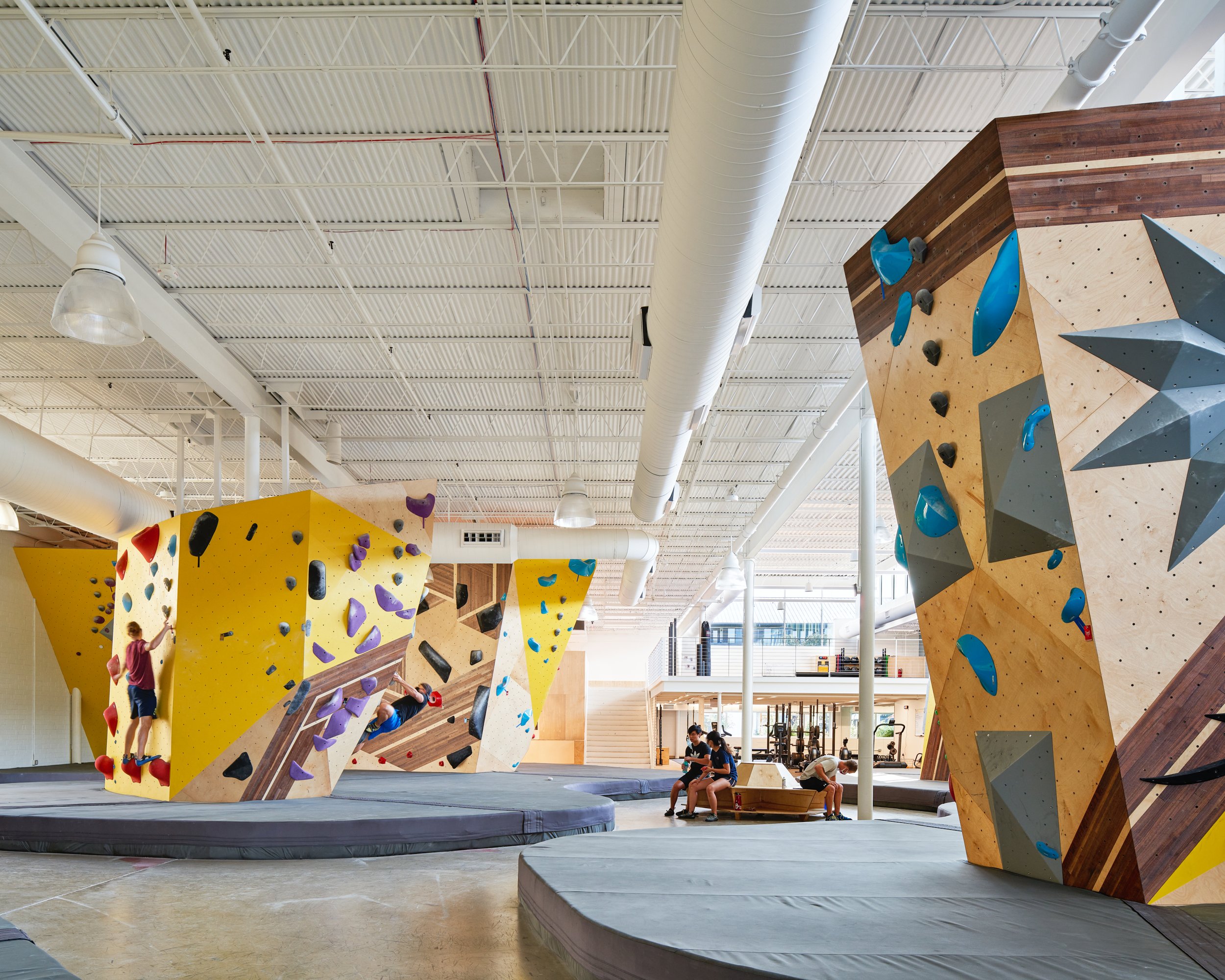 A central view of Crux Central’s main hall, where angular climbing walls, daylight from clerestory windows, and built-in seating define the gym’s open, communal atmosphere.