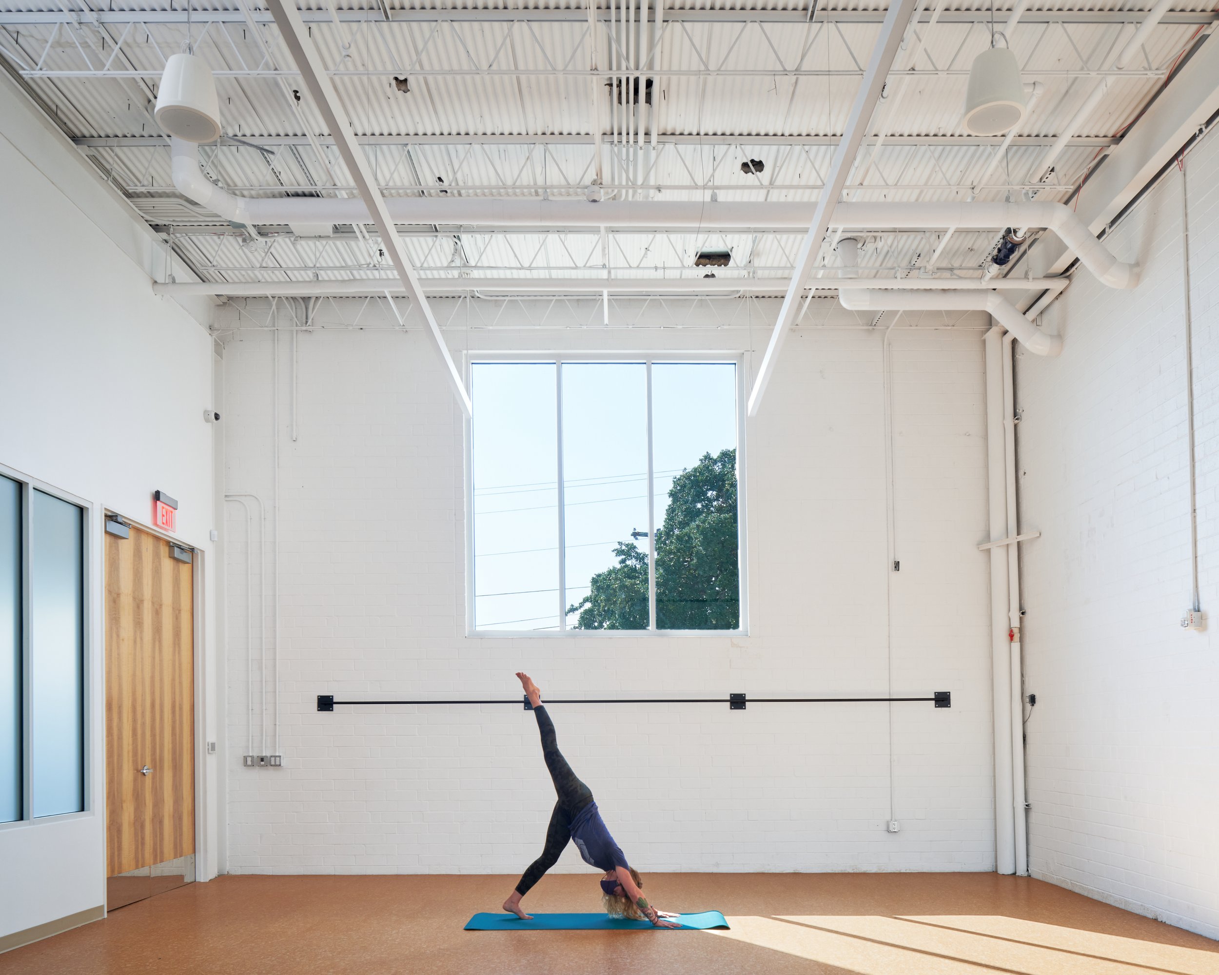 A serene yoga studio framed in white-painted brick and exposed structure, with soft daylight entering through a large window—offering a moment of quiet contrast within the vibrant energy of the gym.