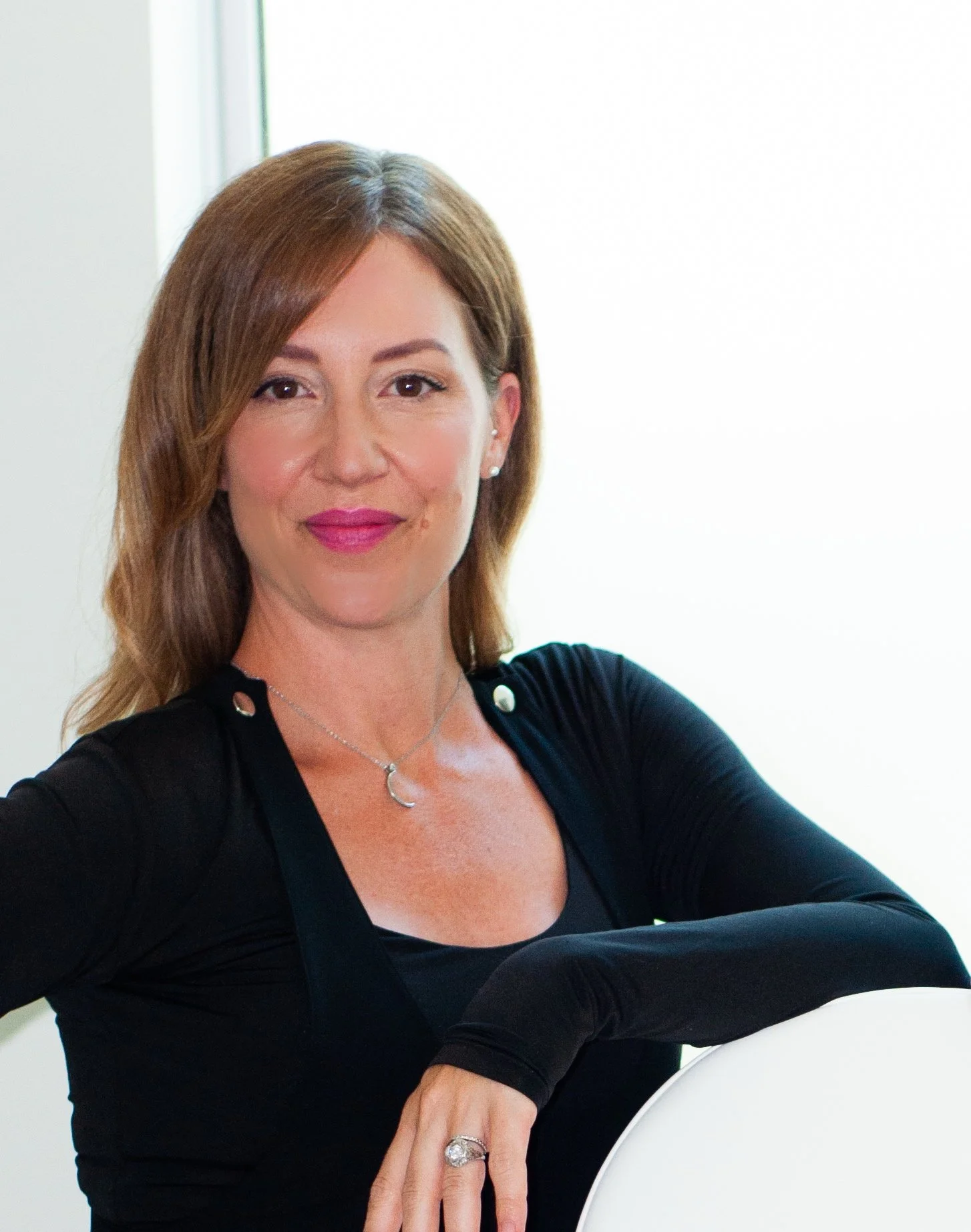A woman from Valere Medical Aestheitcs with shoulder-length brown hair, wearing a black top with silver buttons, a necklace with a crescent moon pendant, and a ring, sitting outdoors near a window with a bright background.
