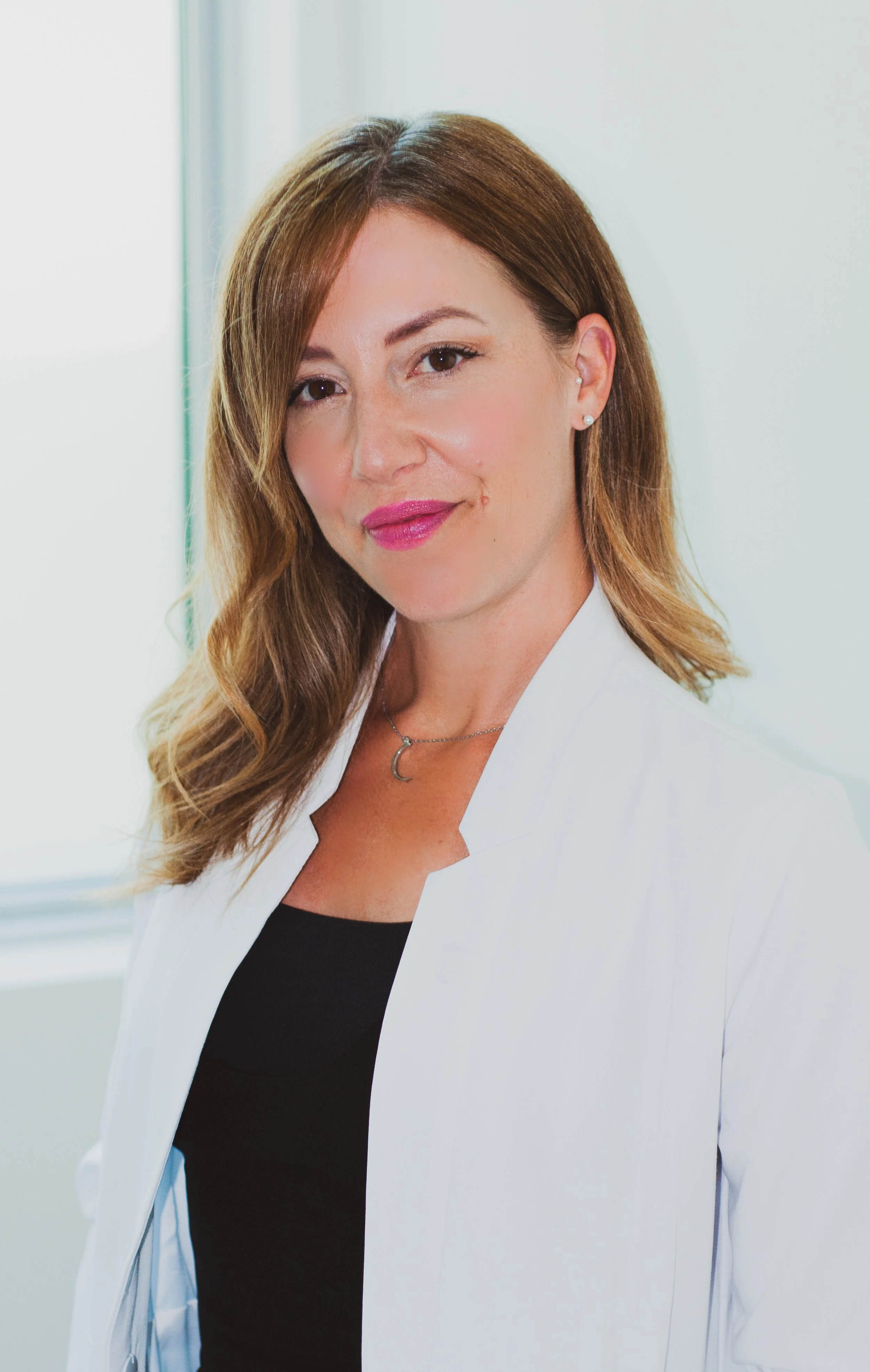 Jennifer Murphy, RN - A nurse from Valere Medical Aesthetics with brown, wavy hair and light skin, wearing a white coat and a black top, standing indoors near a window with natural light.