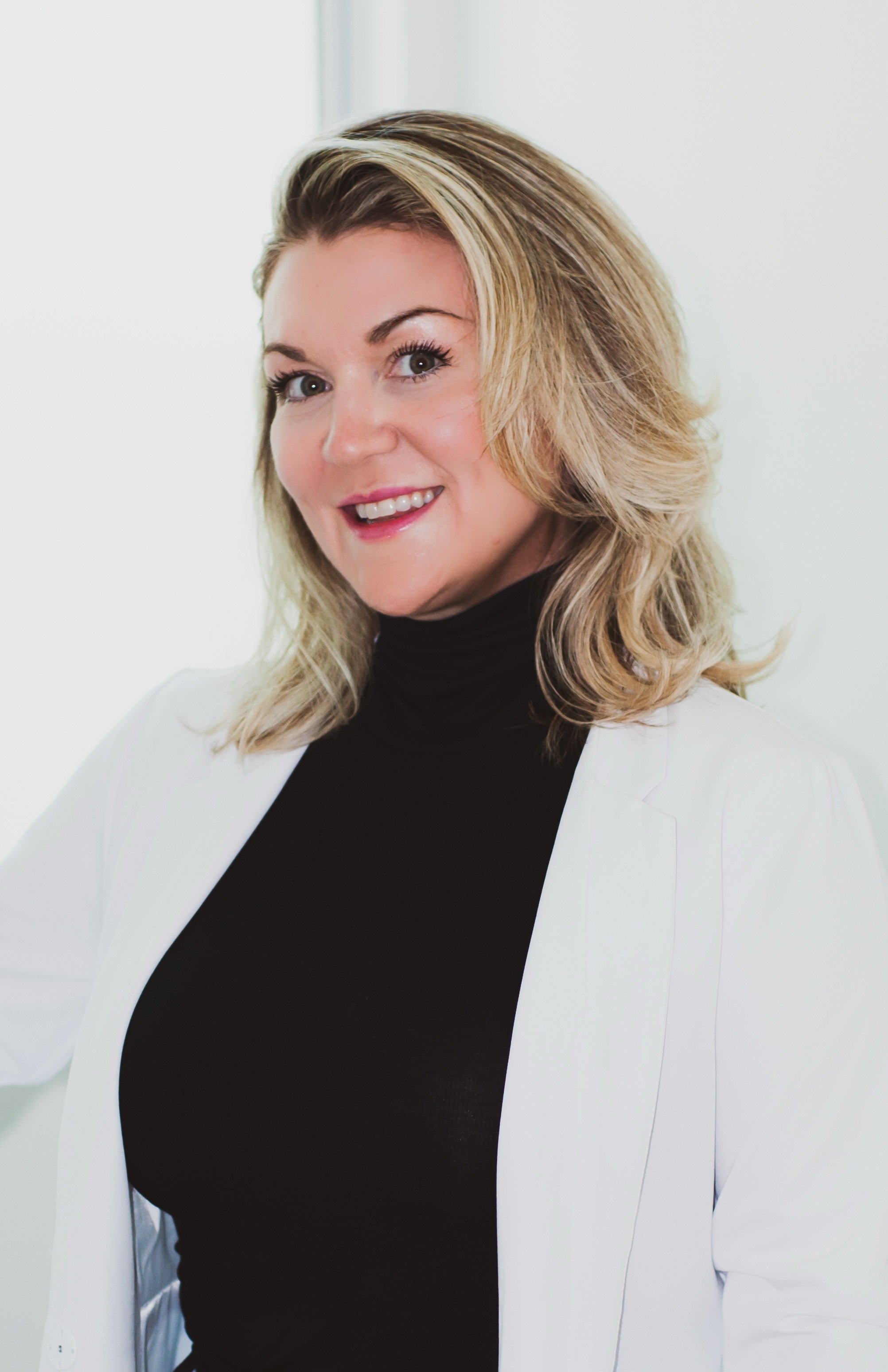 Sara shanks, a valere medical aesthetics nurse is A woman with shoulder-length blonde hair, wearing a black turtleneck and white blazer, smiling at the camera.
