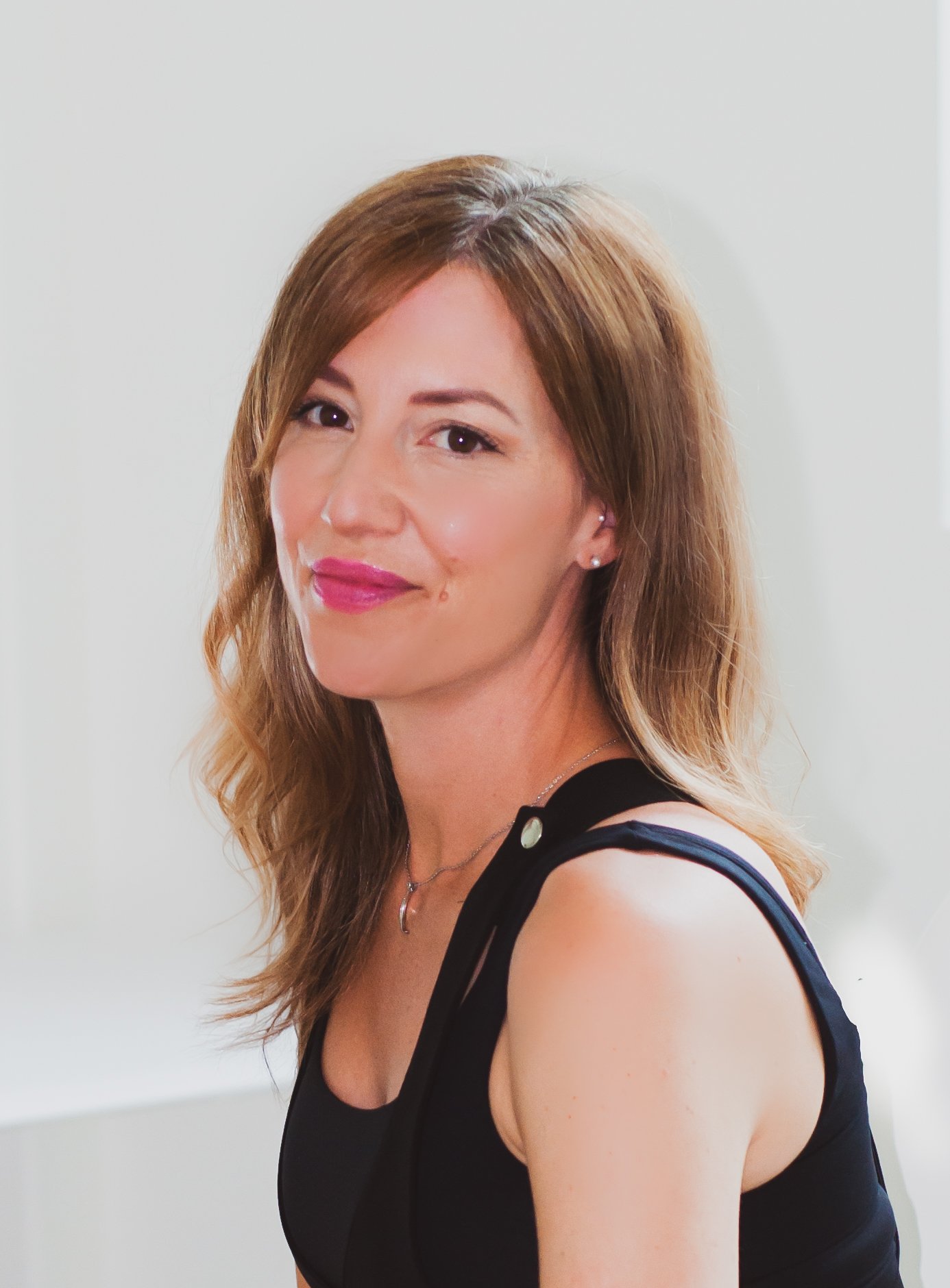 An image of Jen Murphy, a Nurse at Valere Aesthetics. A woman with long, light brown hair, wearing a black sleeveless top, smiling and looking at the camera, standing in a bright room.