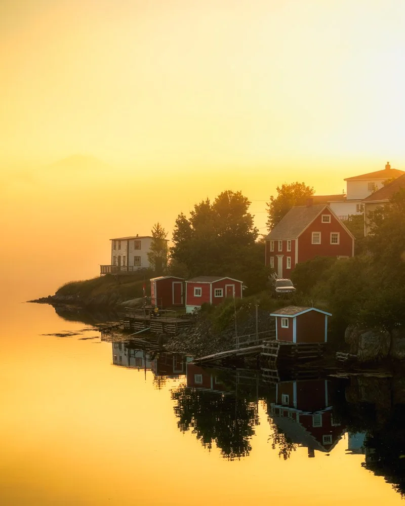 A golden sunrise illuminates the fog and water surrounding Salt Box houses at Buring Newfoundland
