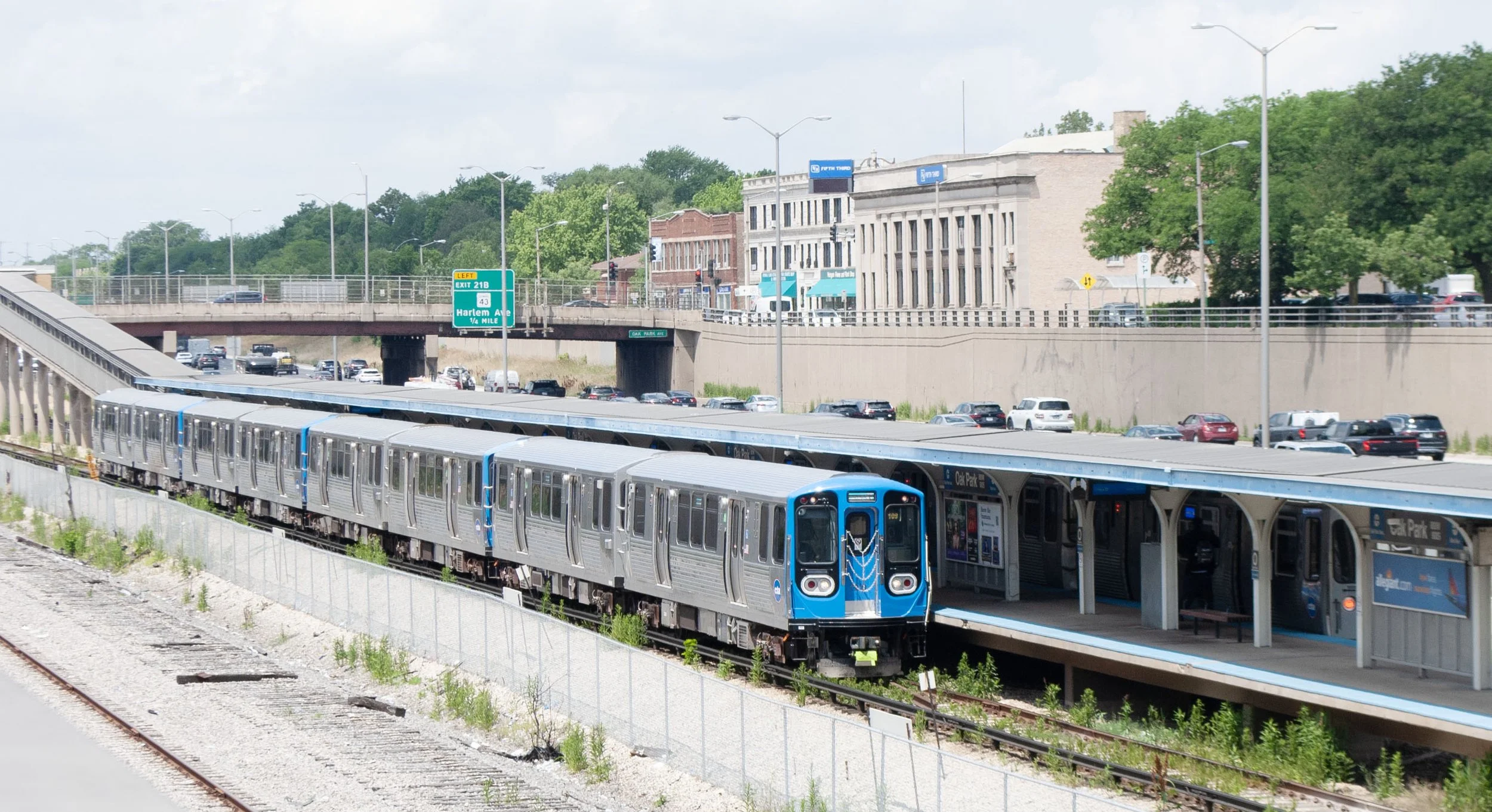 A Blue Line train pulls into Oak Park station with I-290 traffic in the background.