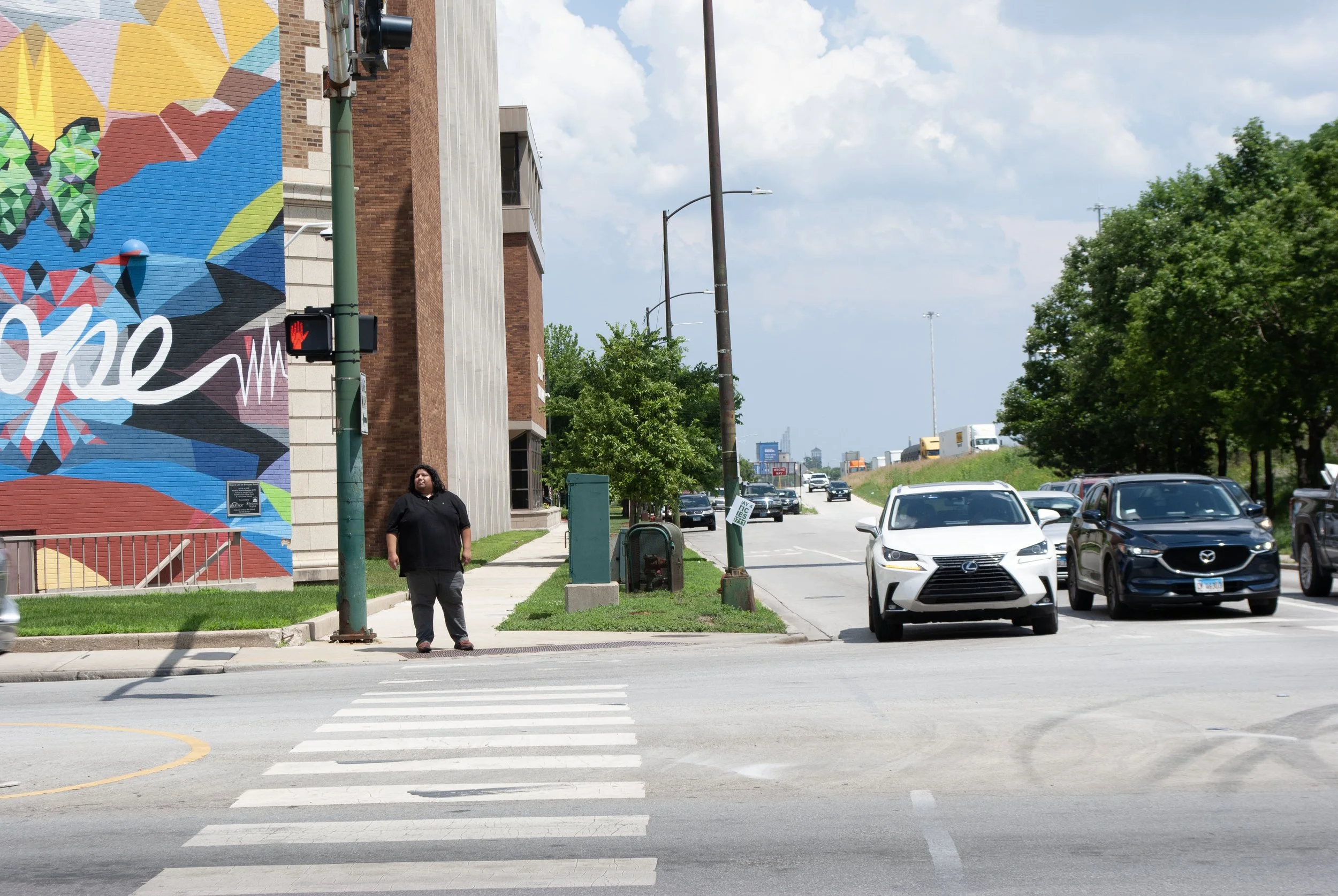 A pedestrian waits to cross the street in front of Loretto Hospital at the I- 290 westbound offramp to Central Avenue. A colorful mural is on a building behind the pedestrian.