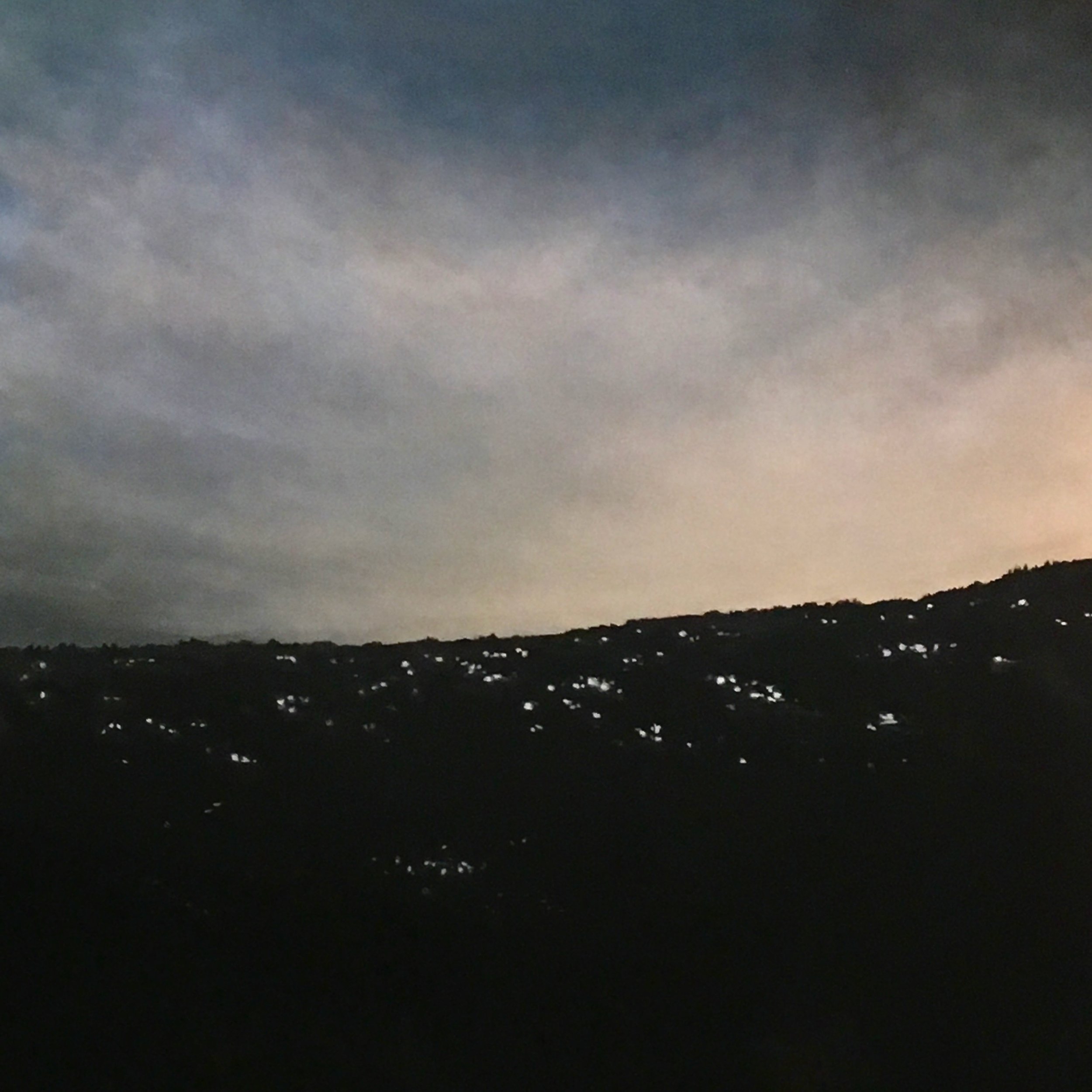 Nighttime landscape with a dark silhouette of hills and scattered lights from houses under a cloudy sky