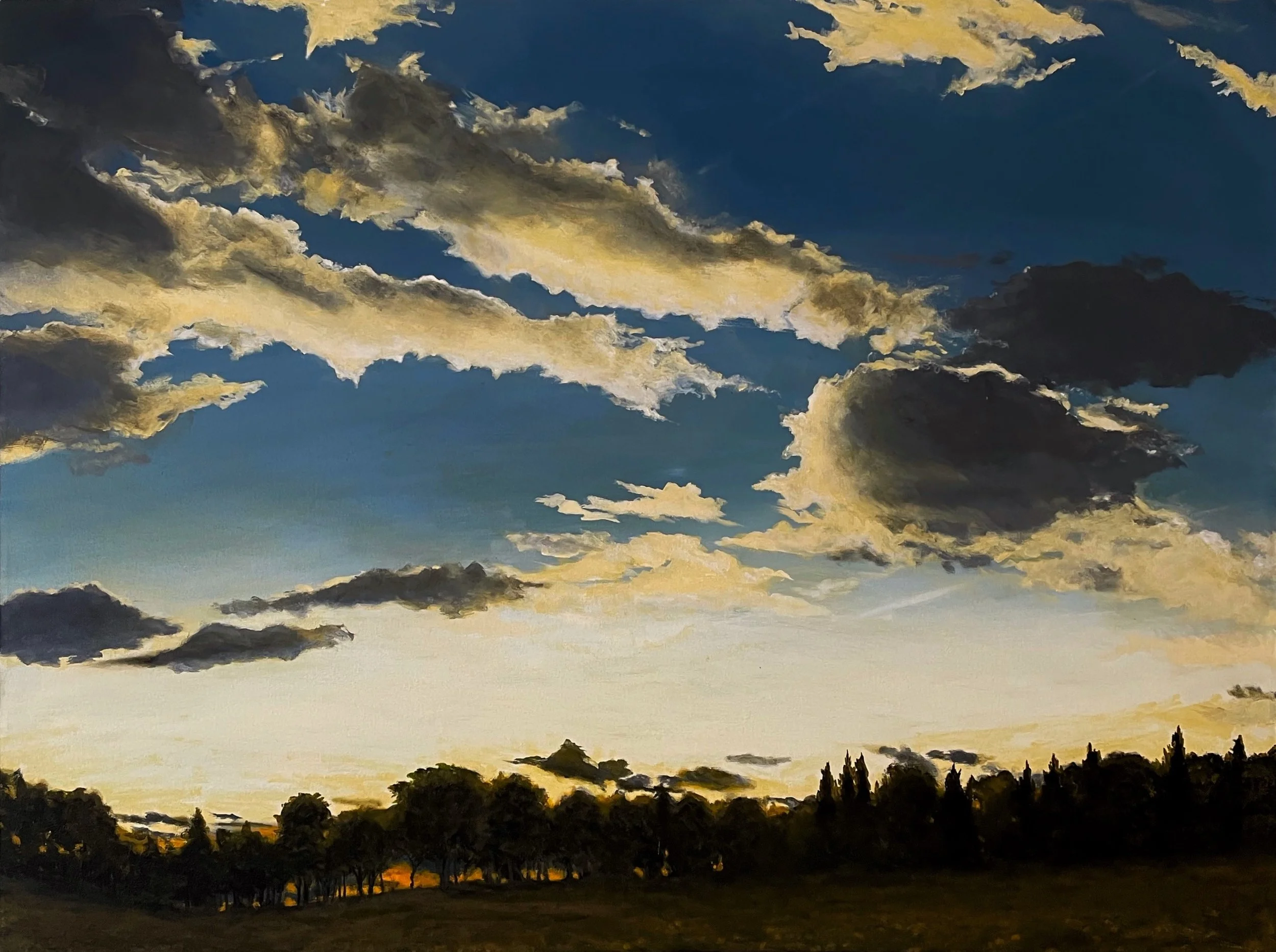 A sunset sky with dark and light clouds above a silhouette of trees and fields.