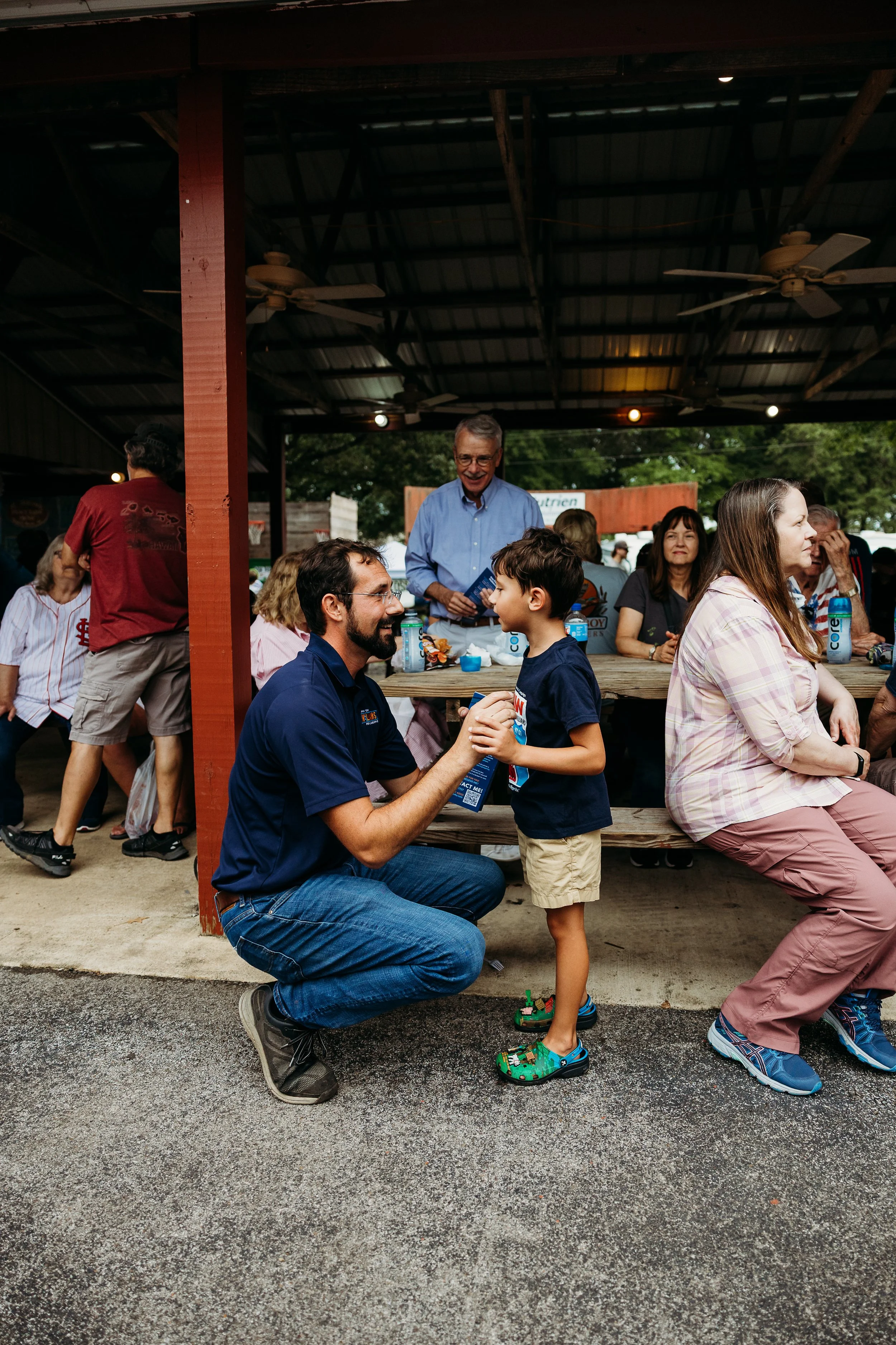 People gather under a covered outdoor area, sitting at picnic tables, engaging in conversations, with a man kneeling and holding hands with a young boy, both smiling. The scene includes various individuals, some standing and some seated, with trees and a fence visible in the background.