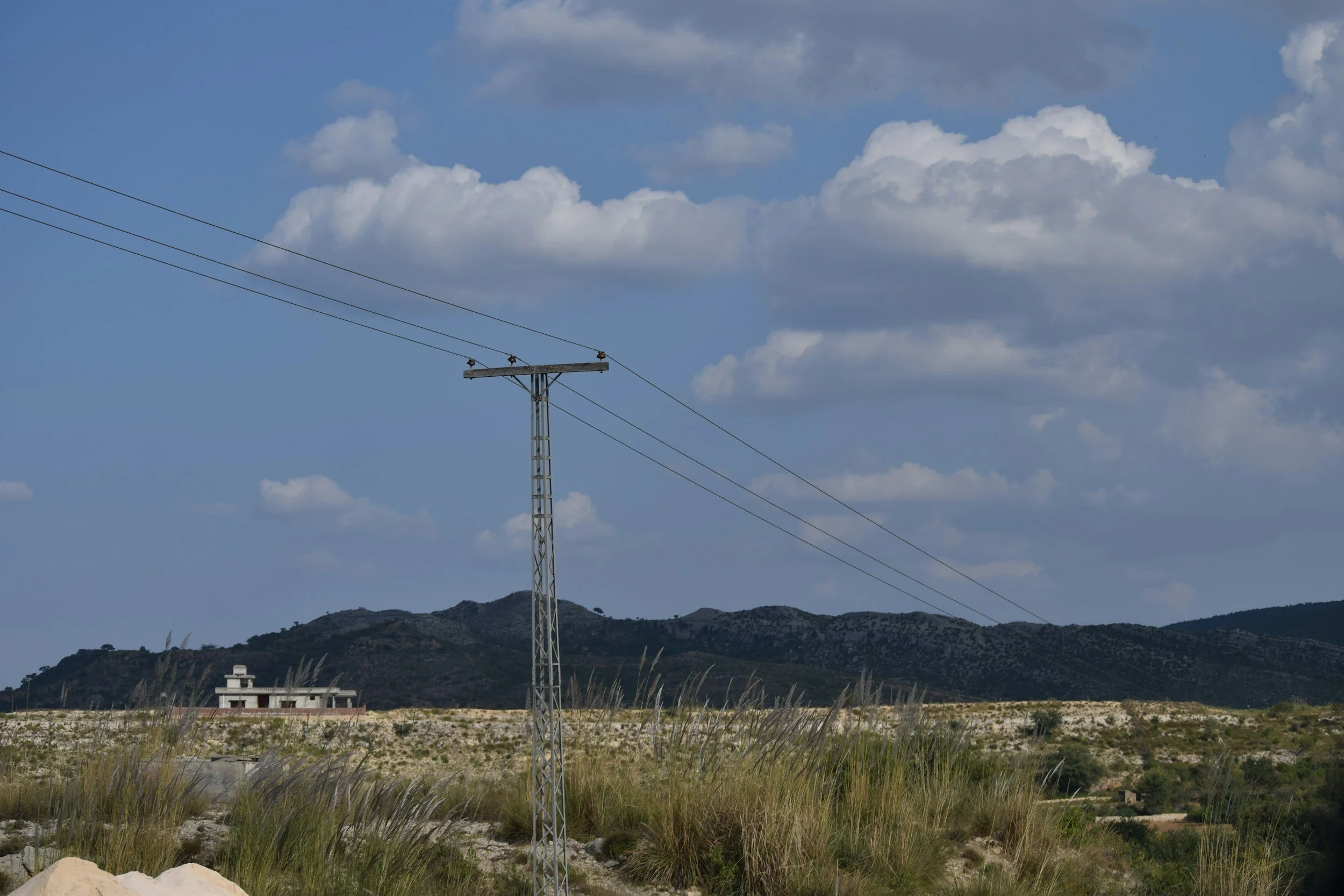 A utility pole with power lines in a rural landscape, with mountains in the background and a partly cloudy sky.