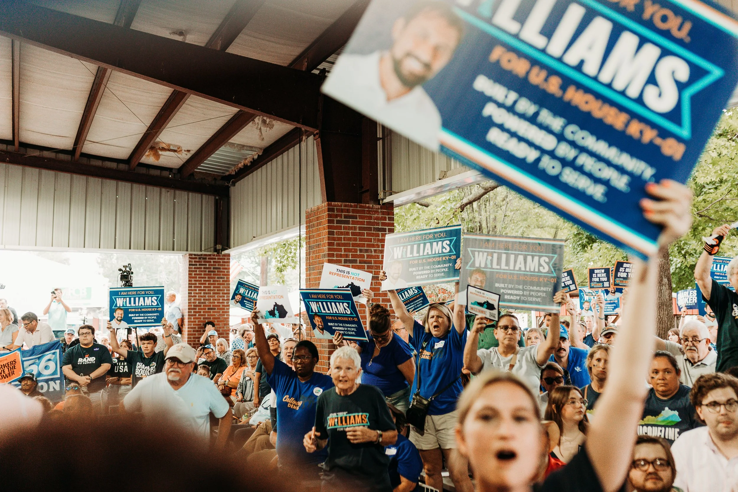 A crowd of people gathered at a political rally holding signs for a candidate named Williams. The signs display the candidate's name and campaign slogan, with some signs stating "Built by the community, powered by people, ready to serve." The rally appears to be outdoors, with a brick building and metal roof structure visible in the background.