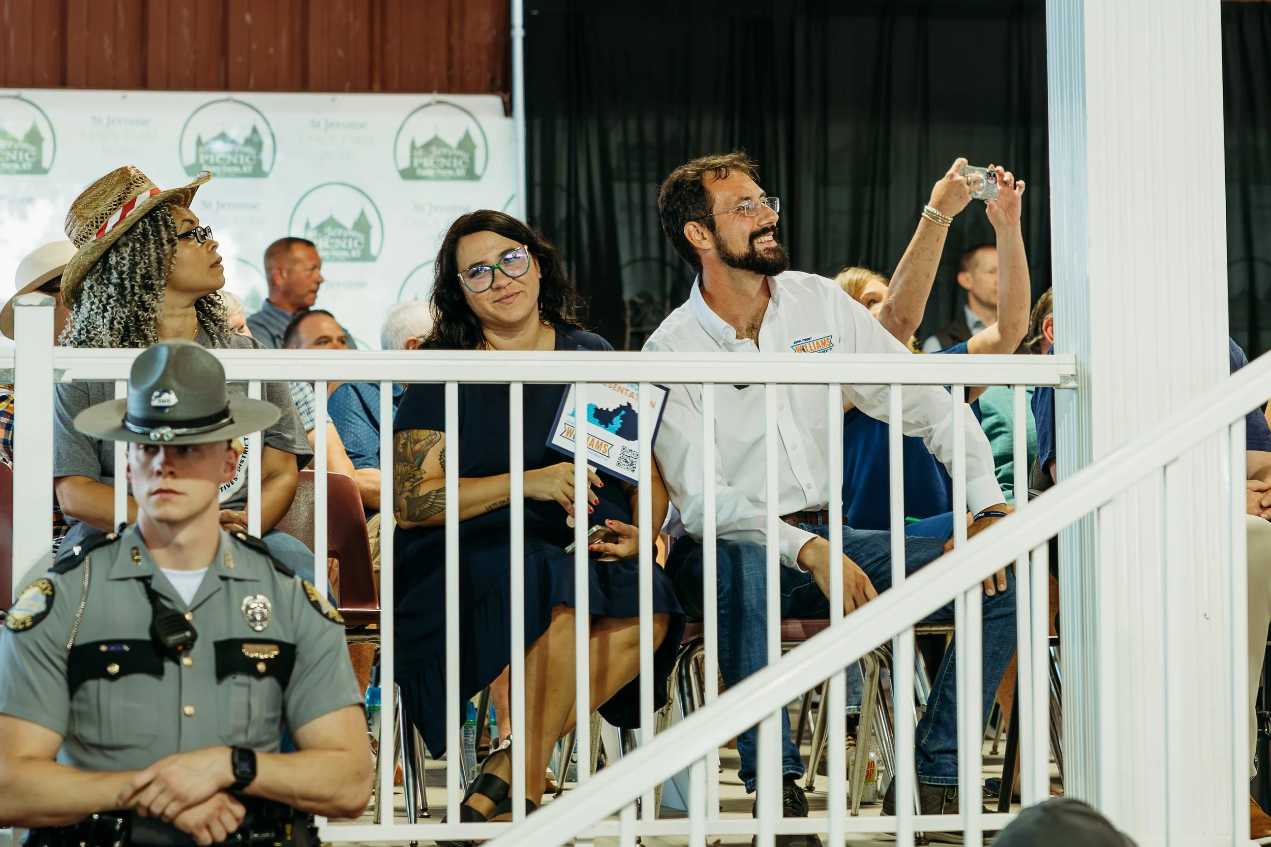People seated in an auditorium, including a woman wearing glasses and a man with a beard taking a photo, a woman with black hair and glasses, a police officer, and others, attending an event with a backdrop displaying logos and text.