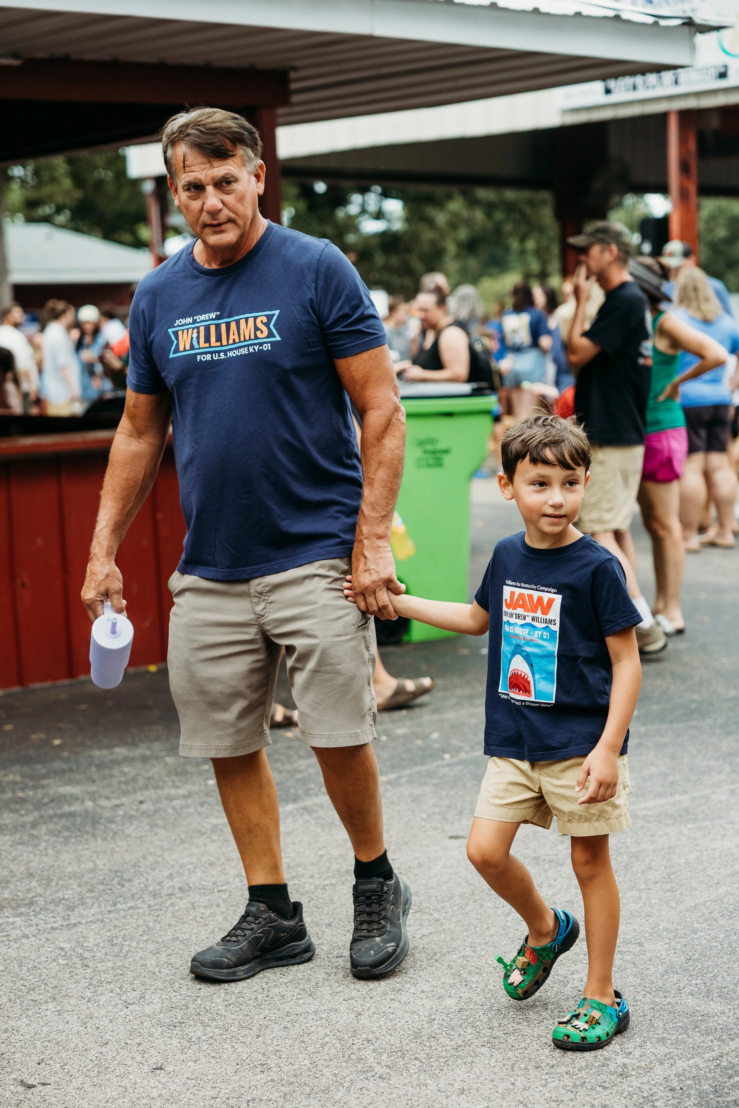 A man and a young boy walking hand in hand at an outdoor event. The man is wearing a blue t-shirt with a political campaign slogan, khaki shorts, and black shoes. The boy is wearing a navy blue t-shirt with a 'Jaws' graphic, beige shorts, and colorful sandals. There are people in the background, some talking and others waiting in line.