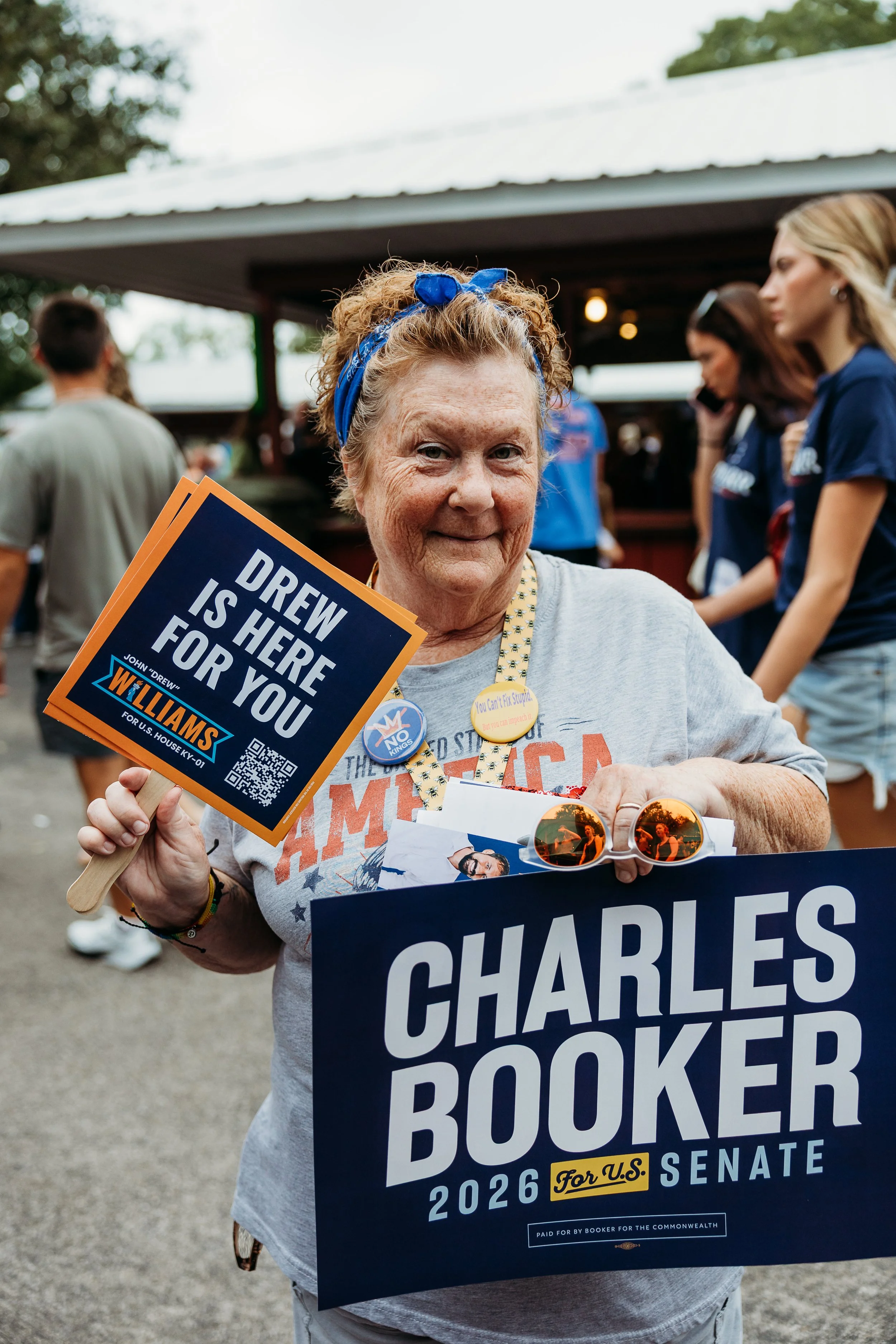Older woman with curly hair wearing a blue headband holding a political sign supporting Charles Booker for U.S. Senate, surrounded by other people at an outdoor event.
