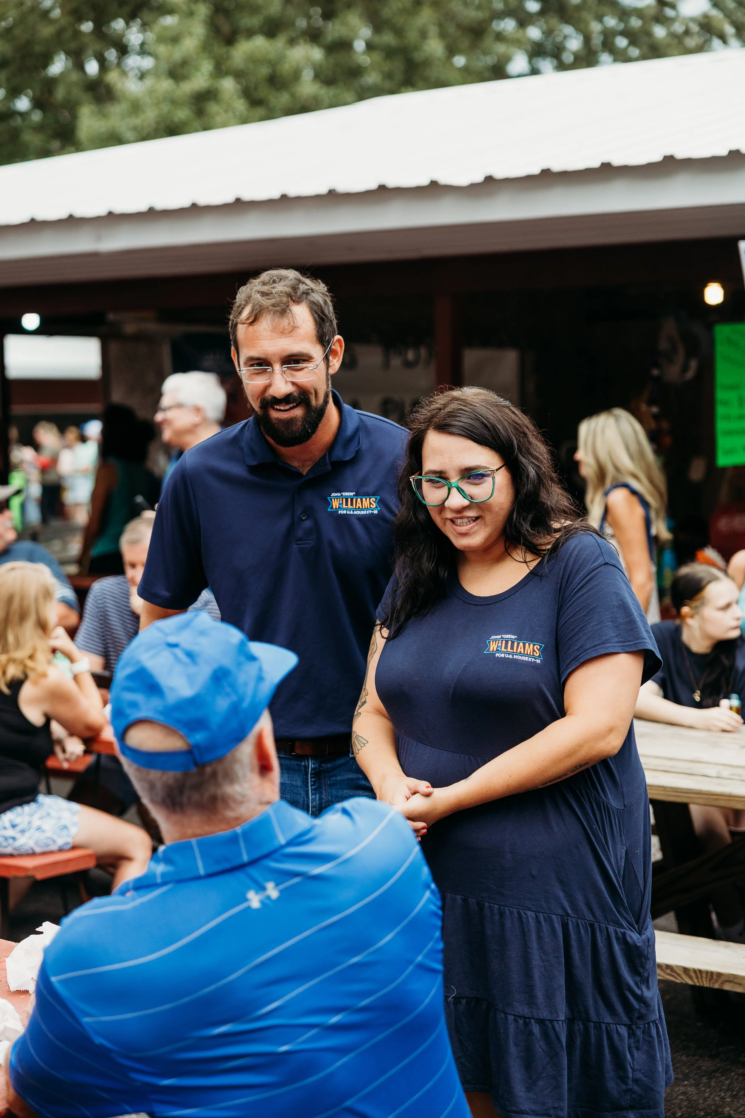 A man with glasses and a beard wearing a blue campaign shirt and a woman with glasses and long dark hair wearing a blue dress speak with a child wearing a blue hat in an outdoor setting with a crowd and trees in the background.