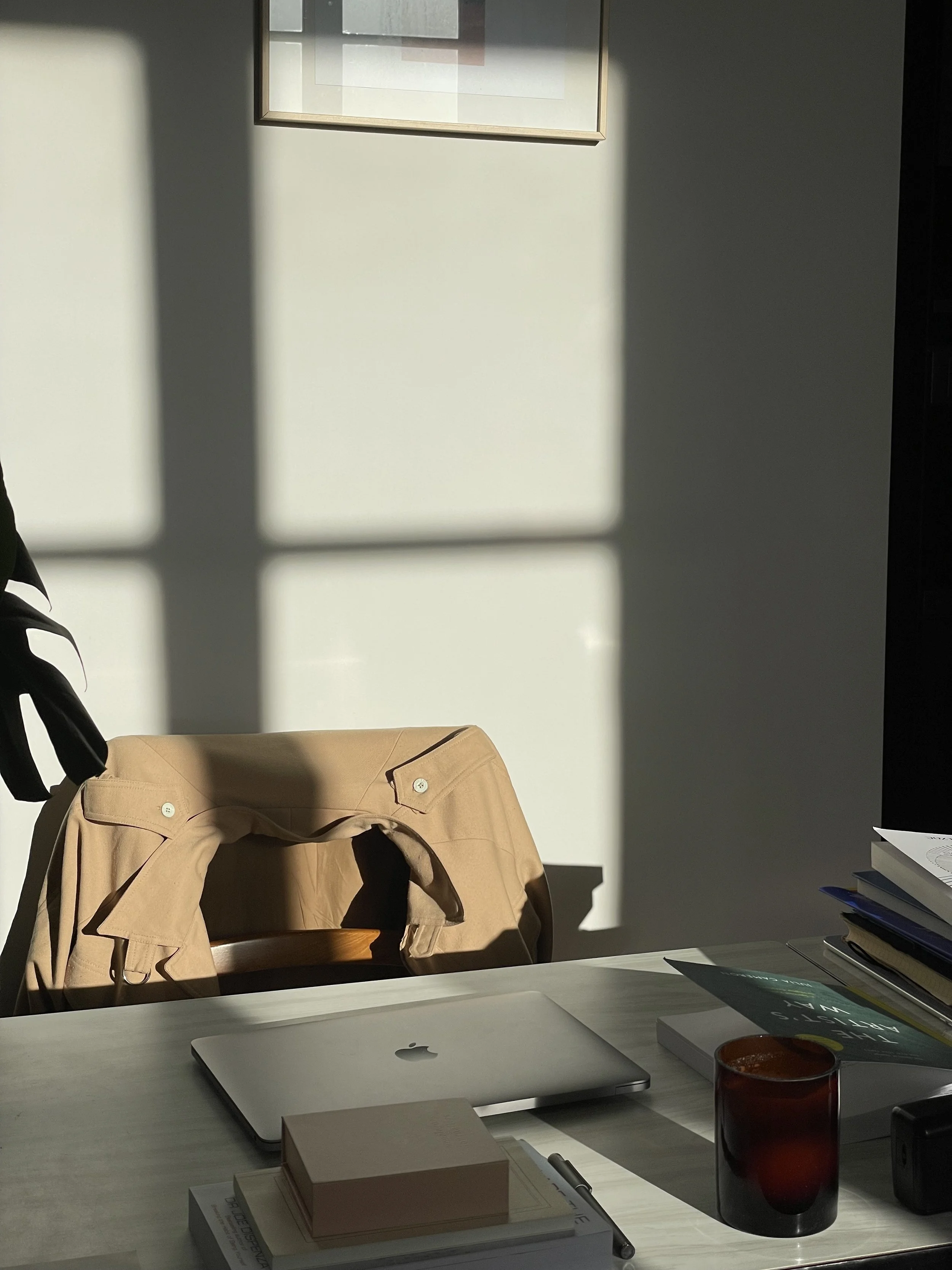 Sunlight casting shadows through a window onto a white desk. On the desk are a closed MacBook, a stack of books including one titled 'The Art of Negotiation,' a red glass, a black electronic device, and some papers and notebooks. A tan blazer is draped over an office chair.