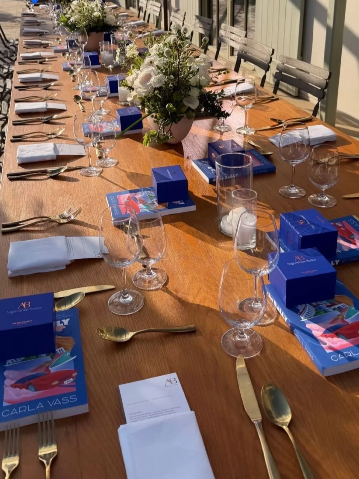 Carla Vass book event dinner in Duck, North Carolina at The Sanderling Resort — a long table set outdoors with wine glasses, white flowers, candles, and blue gift boxes at a Rally launch celebration.