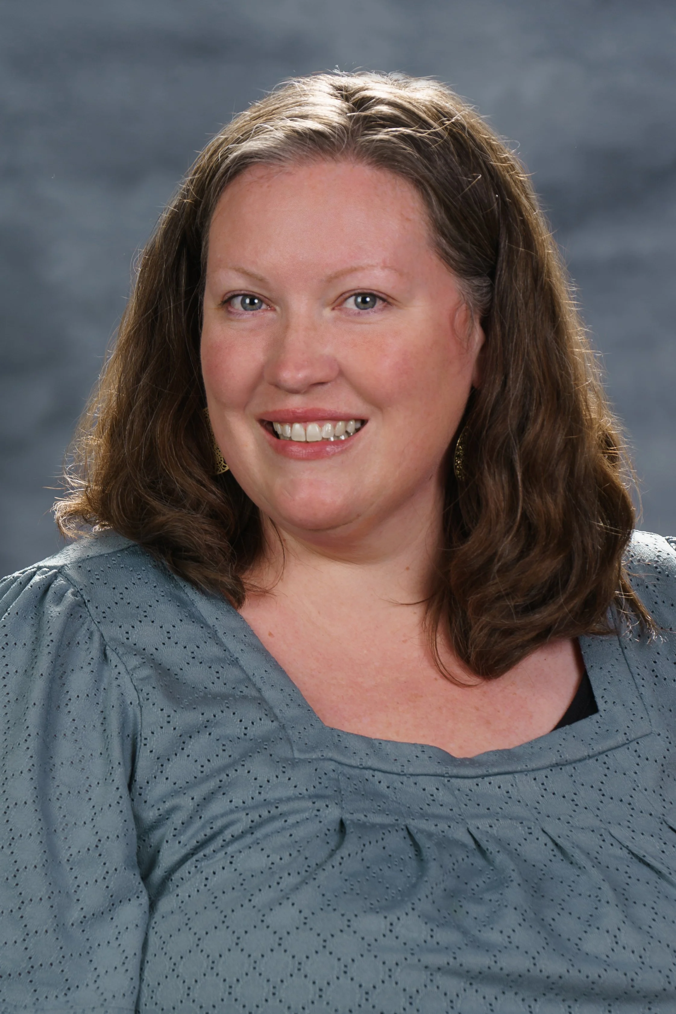 A woman with shoulder-length wavy brown hair, blue eyes, and light skin, smiling, wearing a gray textured blouse with a black top underneath, and earrings, against a gray background.