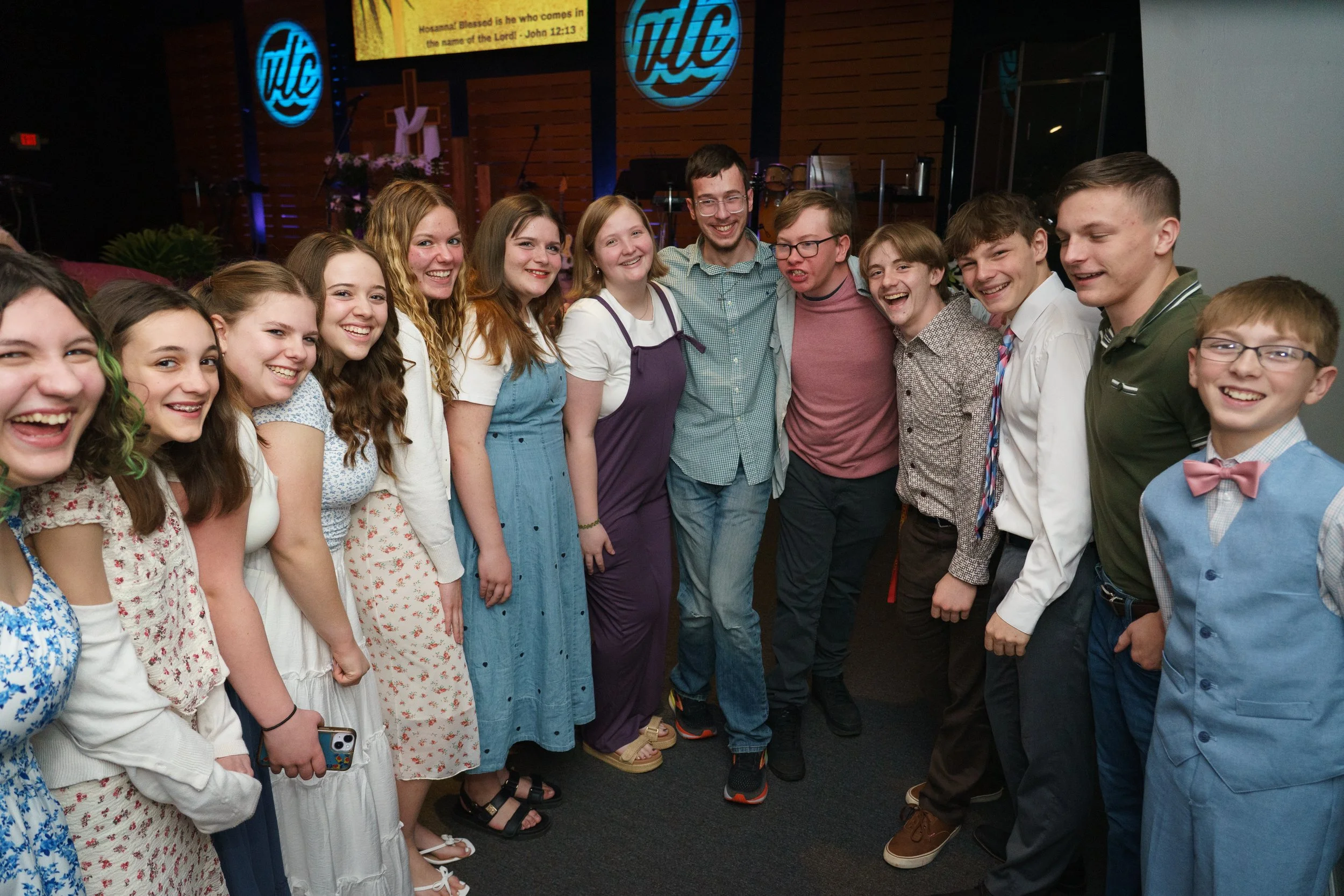 Group of young people, standing together in church, smiling and posing for a group photo, with a church logo and a cross in the background at Victory Life Church near Hudson, OH.
