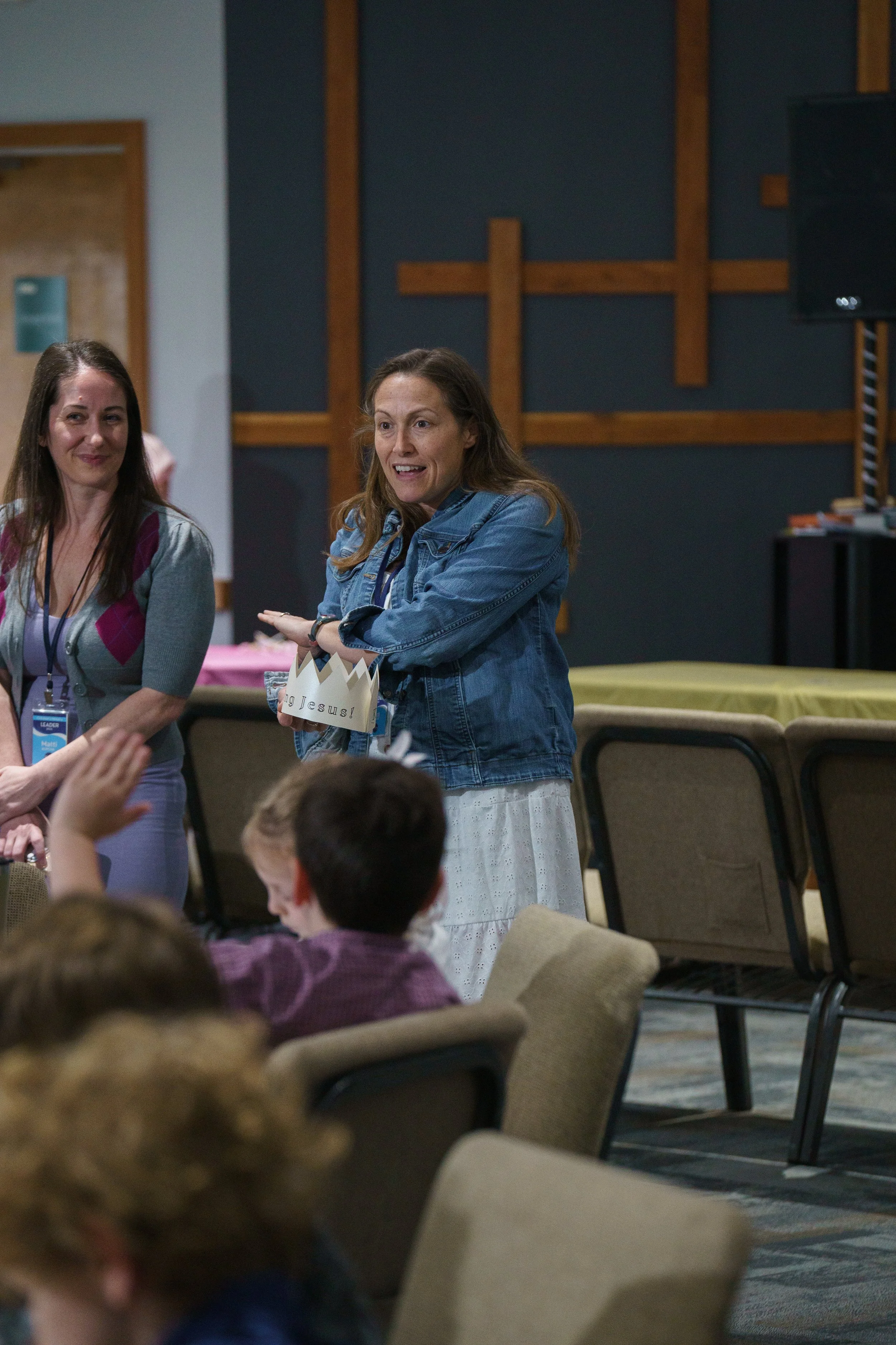 A woman with long brown hair, wearing a white dress, is speaking to a group of children. The woman is holding a paper crown with the words "King Jesus" on it as she teaches at Victory Life Church in Stow, OH, near Streetsboro, OH.
