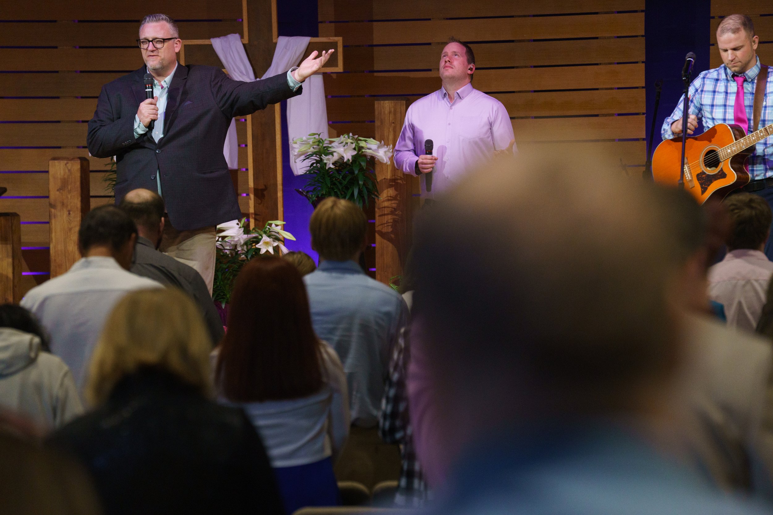 A pastor in a dark suit and glasses speaking into a microphone, a man with a guitar, on a stage with wooden decor and a cross in the background during a church service at Victory Life Church in Stow, OH, near Hudson, OH.