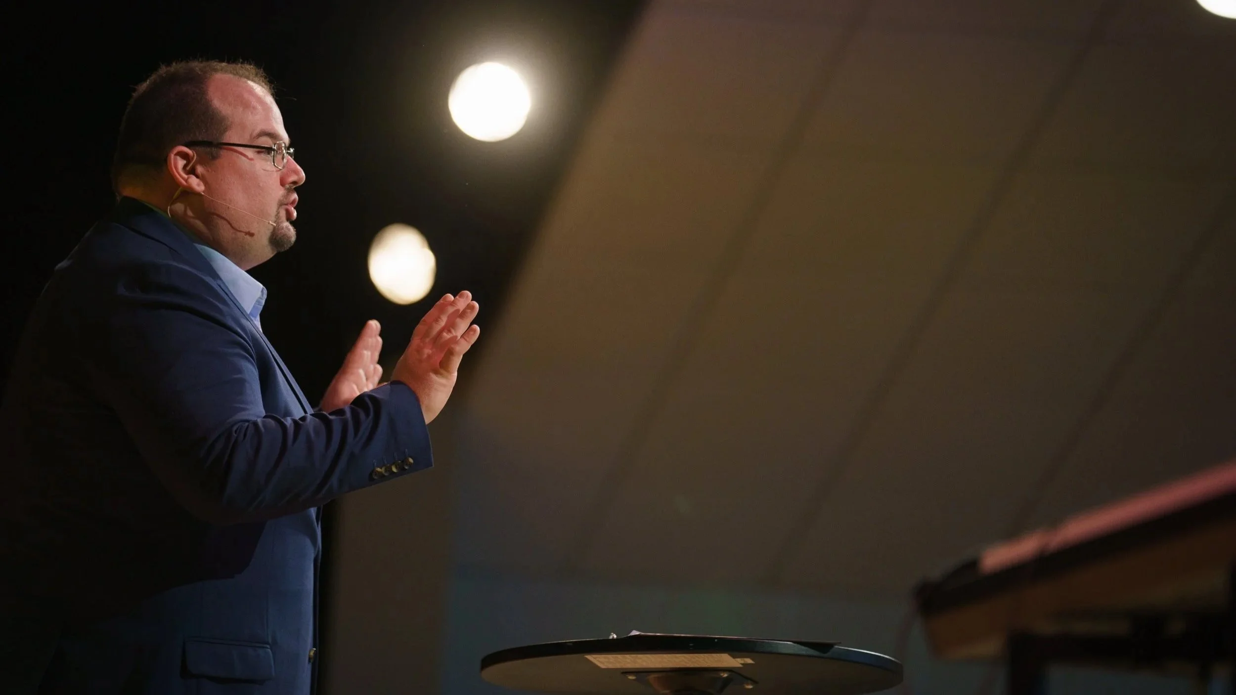 Pastor Matt Skifstad in a blue suit and glasses, with one hand raised, standing in front of a dark background with bright circular lights, is speaking at Victory Life Church in Stow, OH near Streetsboro, OH.