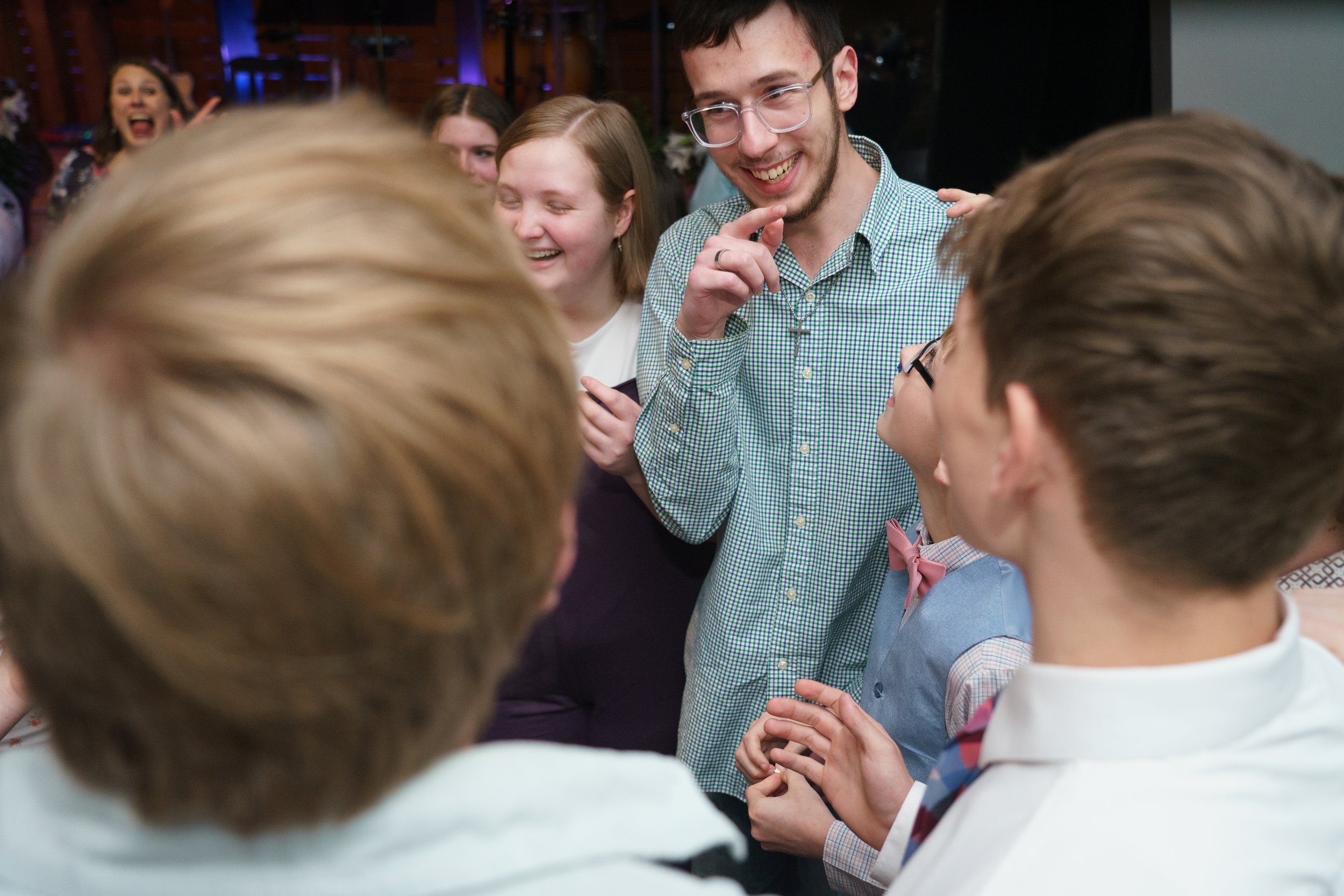 A group youth of gather closely, smiling and engaging in conversation at church. The focus is on a young man with glasses and a checked shirt who is smiling. At Victory Life Church in Stow, OH near Cuyahoga Falls, OH.
