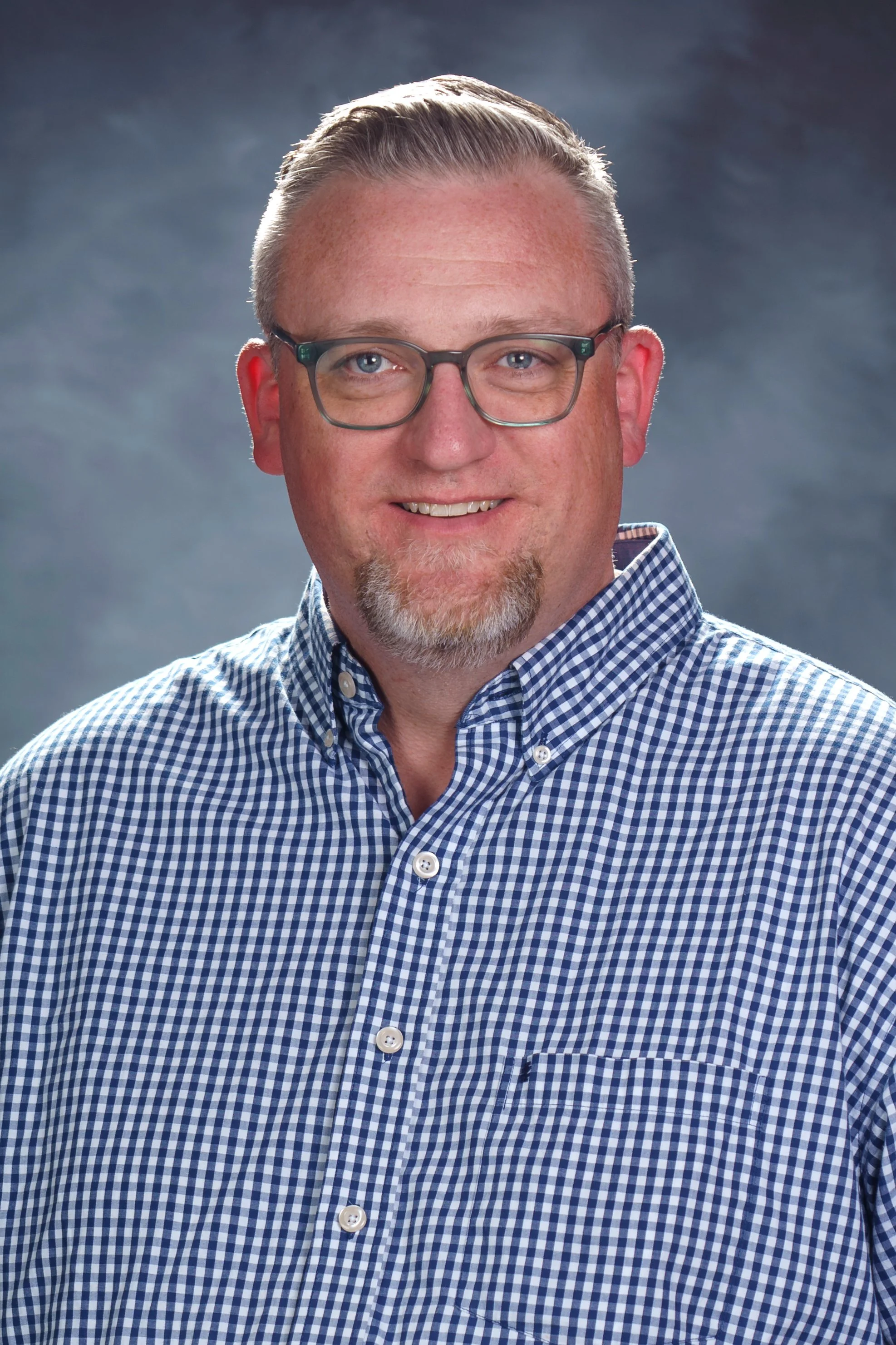 A middle-aged man with short, light-colored hair, glasses, and a goatee, wearing a blue and white checkered shirt, smiling at the camera.