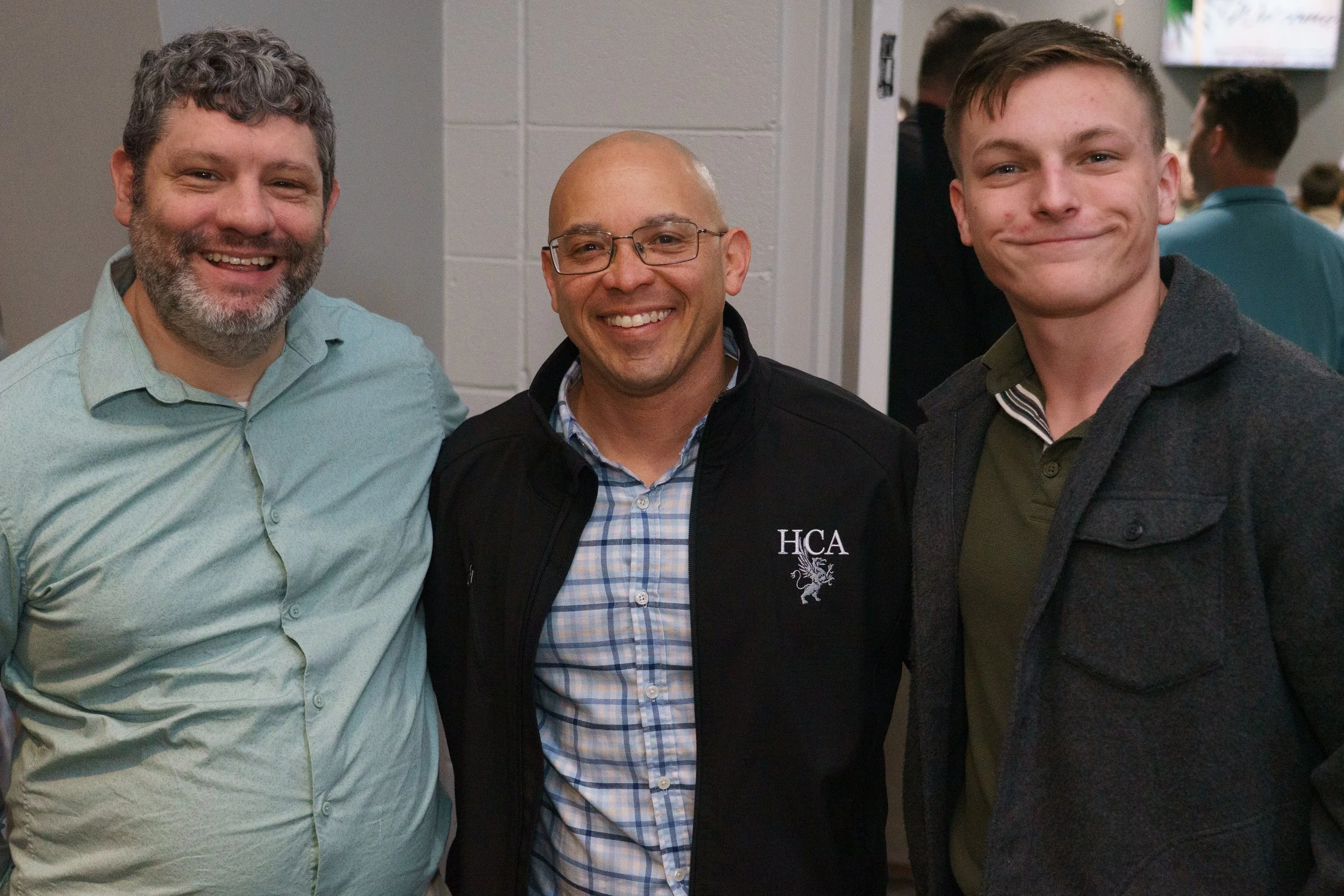 Three men standing and smiling together after a church service at Victory Life Church in Stow, OH, near Hudson, OH with a gray wall behind them and other people in the background.