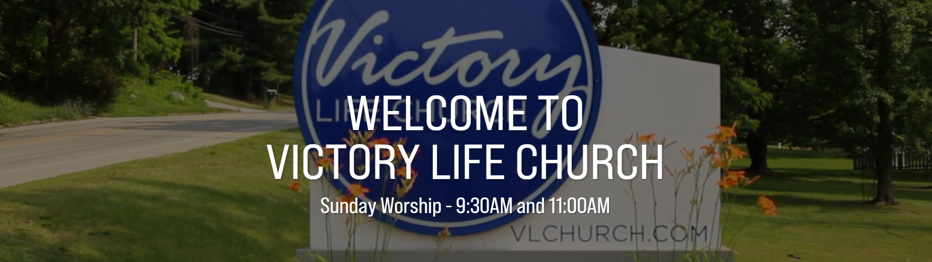 Sign for Victory Life Church with welcoming message and service times, outdoor setting with trees and grass