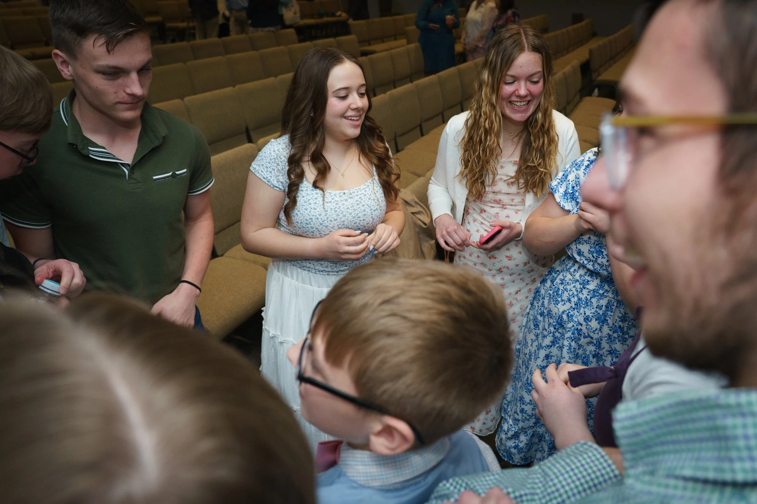 Group of young people gathered in a room, some smiling and talking, with chairs in the background.
