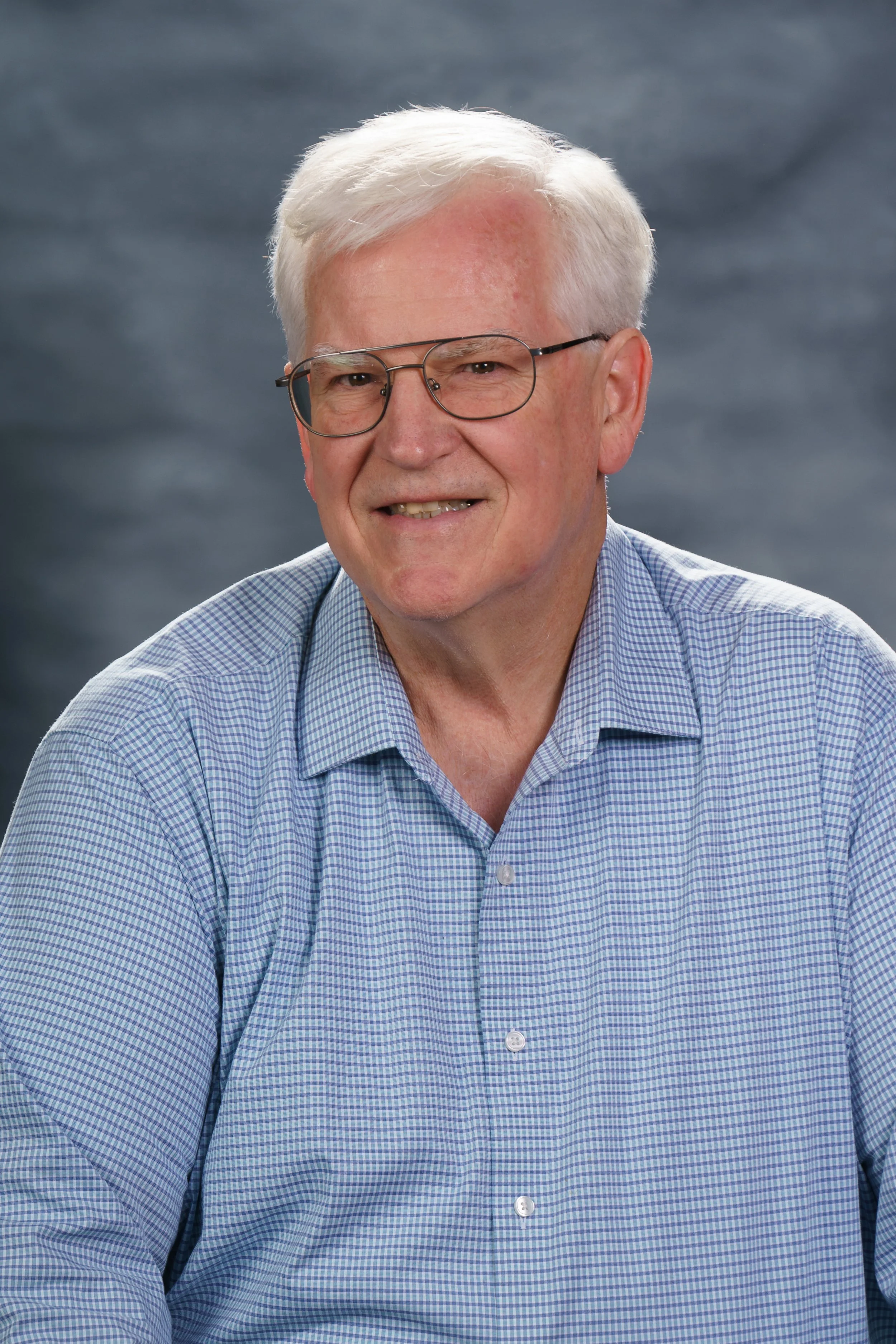 An elderly man with gray hair, glasses, and a light blue checkered shirt smiling against a gray backdrop.