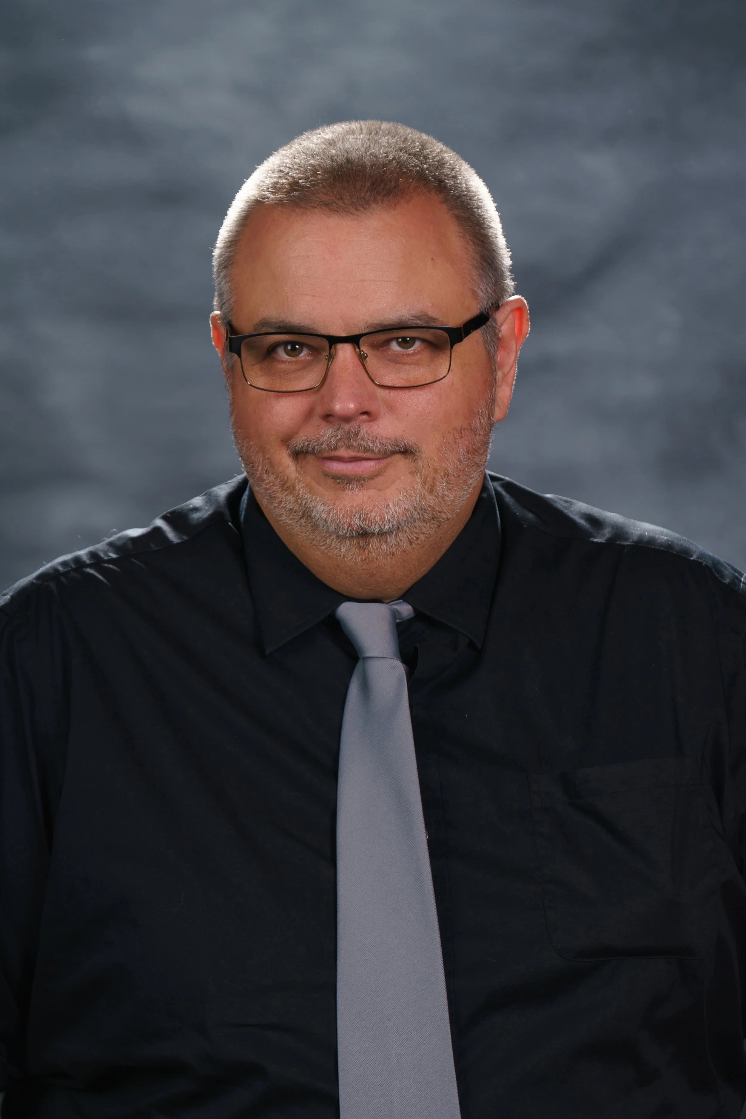A middle-aged man with short gray hair, glasses, and a trimmed beard, dressed in a black shirt and gray tie, posing against a dark, stormy background.