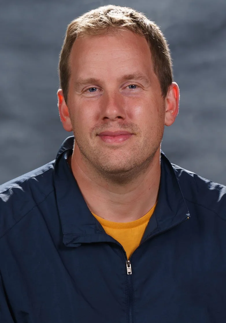 Close-up portrait of a man with short blond hair, blue eyes, and a slight smile, wearing a navy zip-up jacket over a yellow shirt, with a blurred gray background.
