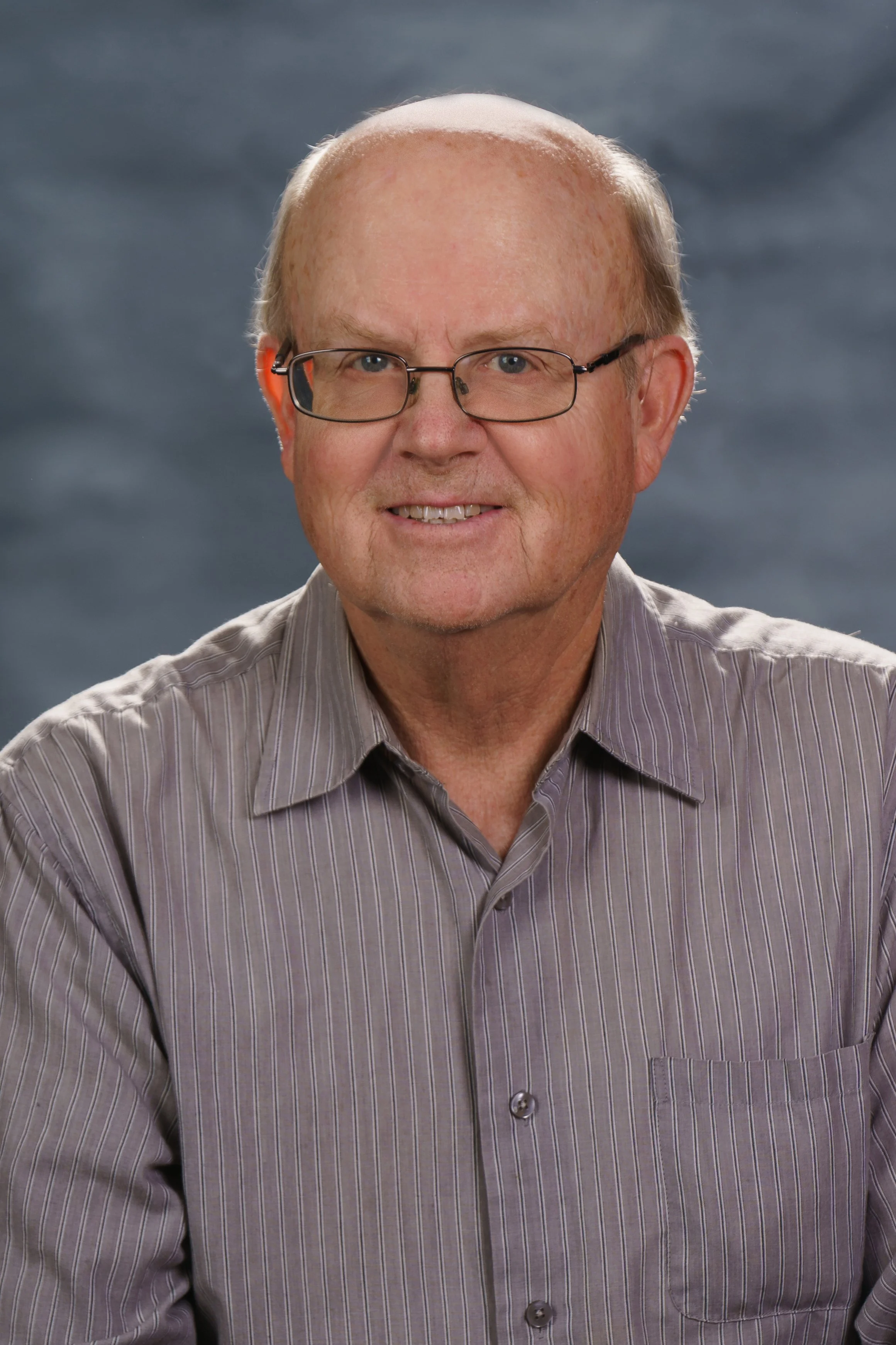A portrait of an older man with glasses, wearing a striped shirt, and smiling at the camera against a dark, cloudy background.