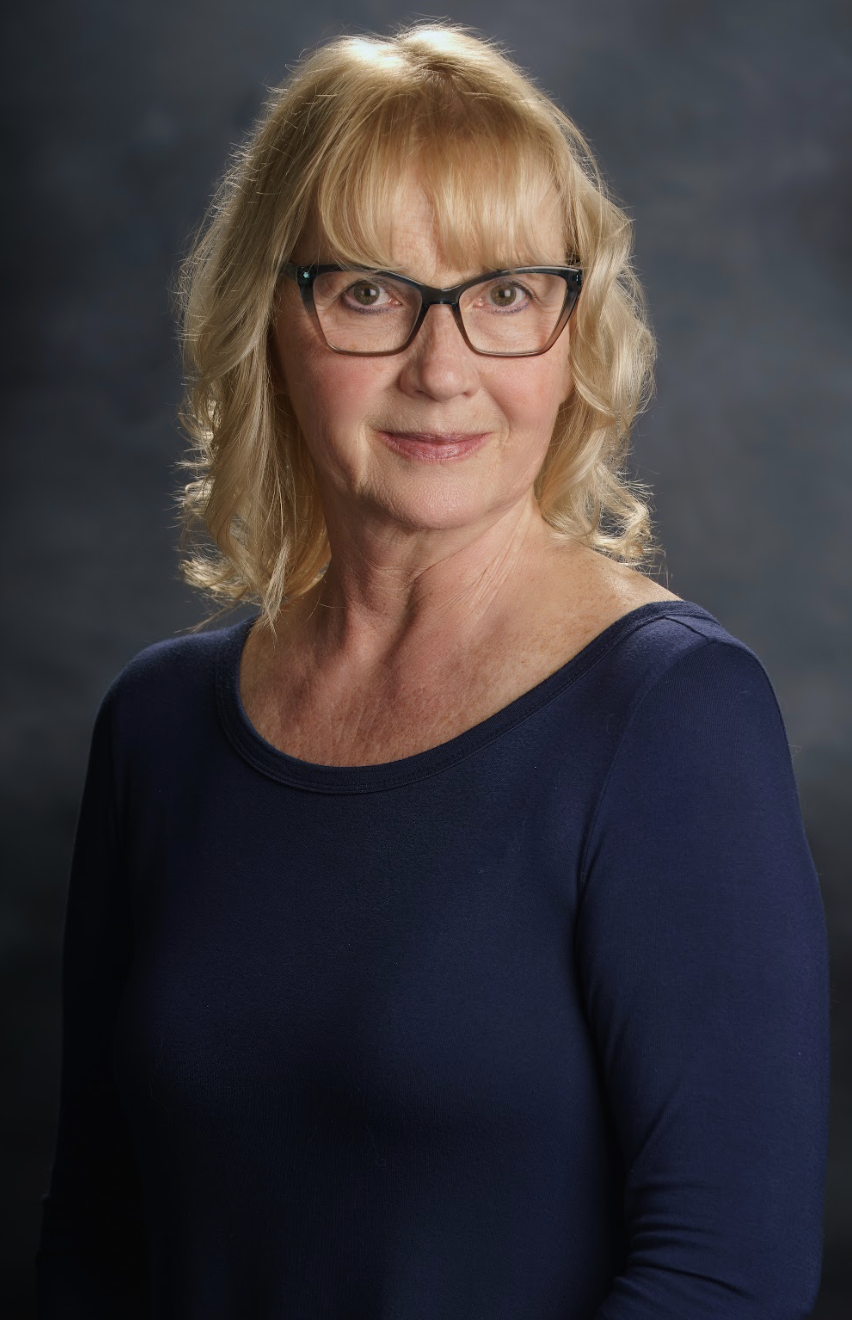 A middle-aged woman with blonde hair, glasses, and a navy blue top, smiling softly against a dark, cloudy background. She is the Children's Program Specialist at Victory Life Church.