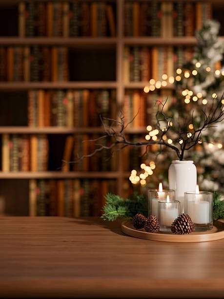 Decorative holiday centerpiece with candles, pinecones, and branches in a white vase on a wooden table in front of a blurred bookshelf and Christmas tree with lights.