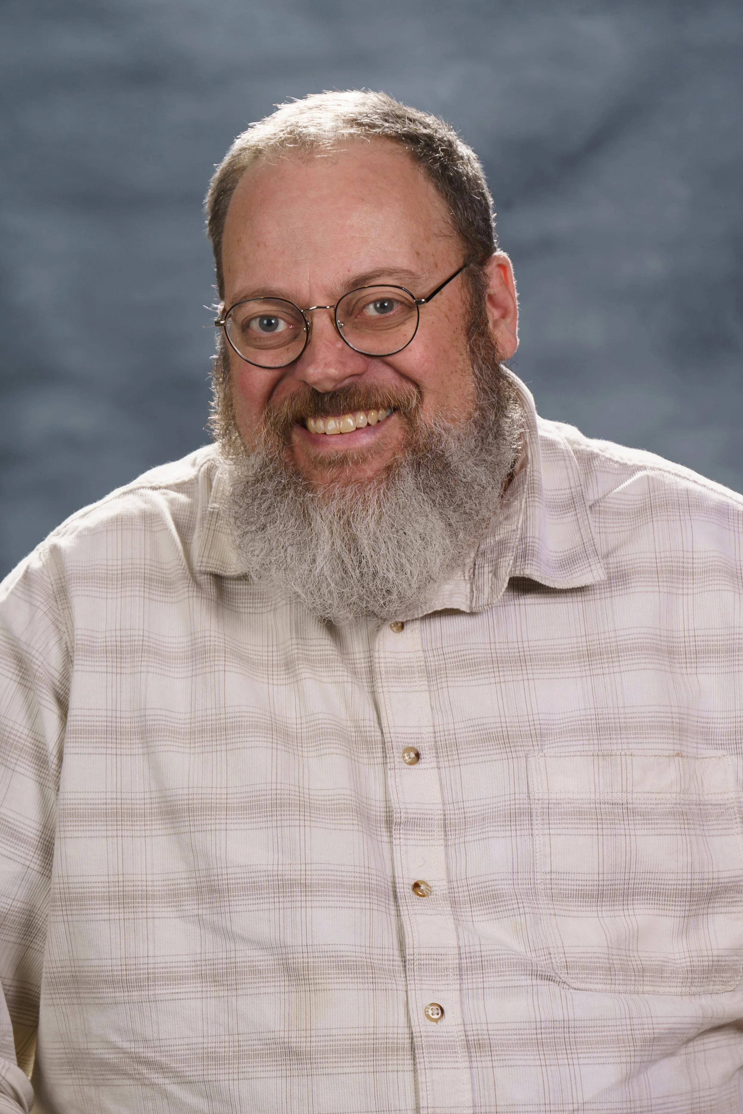 A smiling man with glasses, a full beard, and a checkered shirt against a dark background.