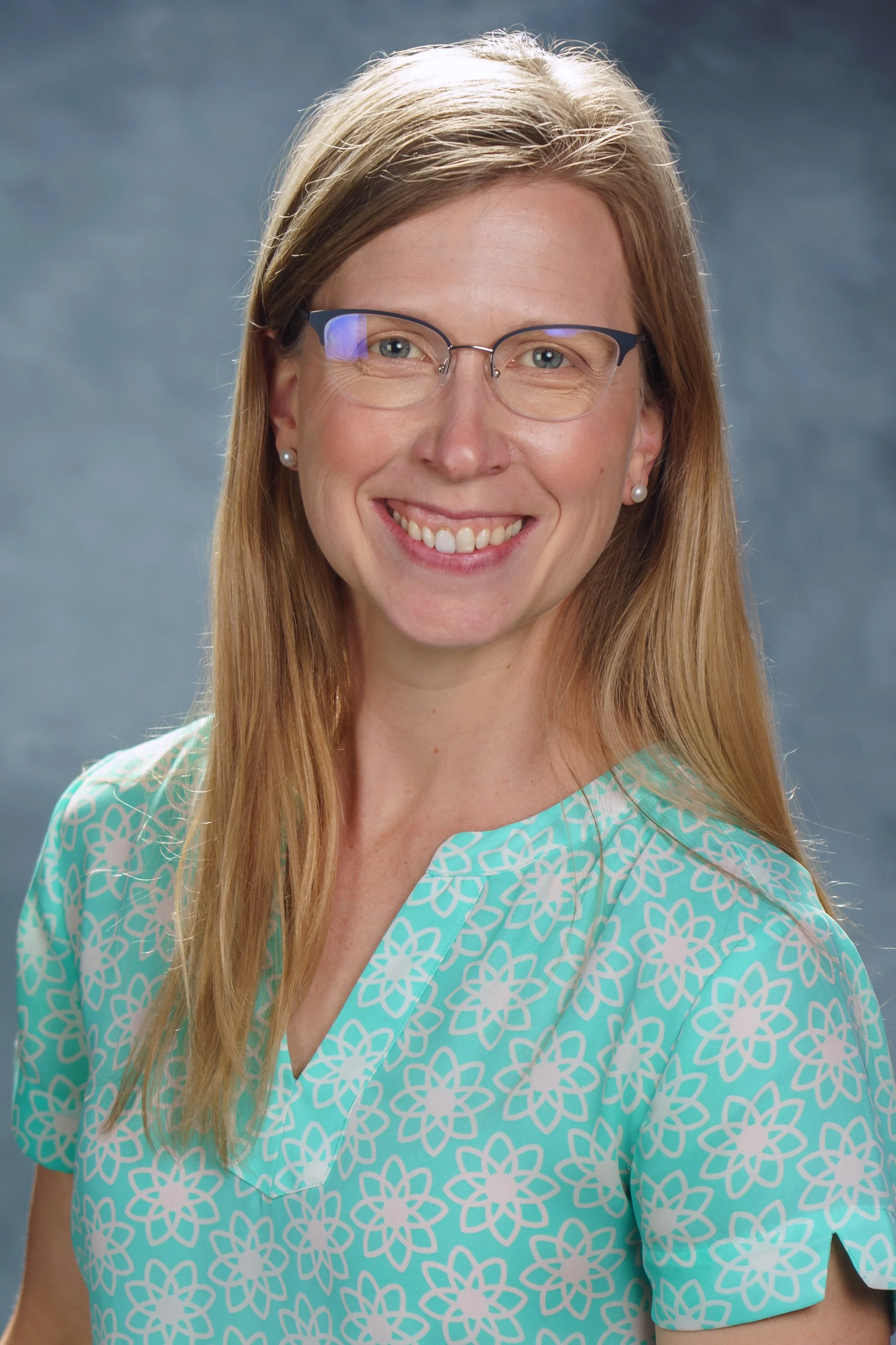 Portrait of a smiling woman with long blonde hair, glasses, and earrings wearing a light blue patterned shirt with white floral designs, against a gray background.