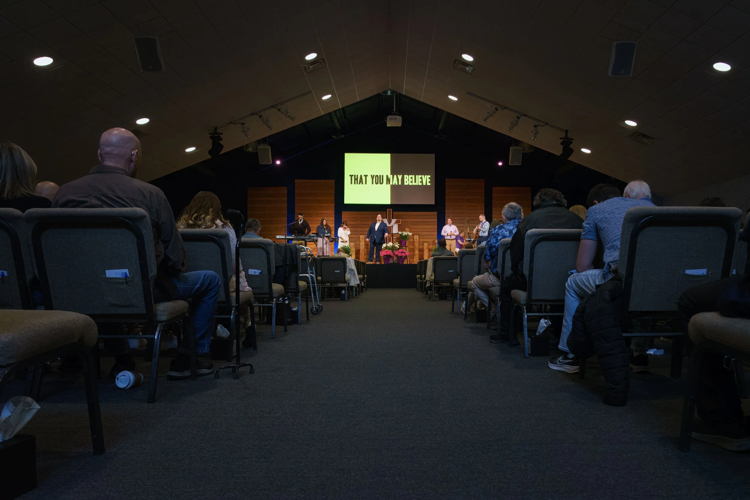Indoor church service at Victory Life Church in Stow, OH with people seated facing a stage. On the stage, a speaker is speaking into a microphone, and a large screen displays the words 'THAT YOU MAY BELIEVE'.