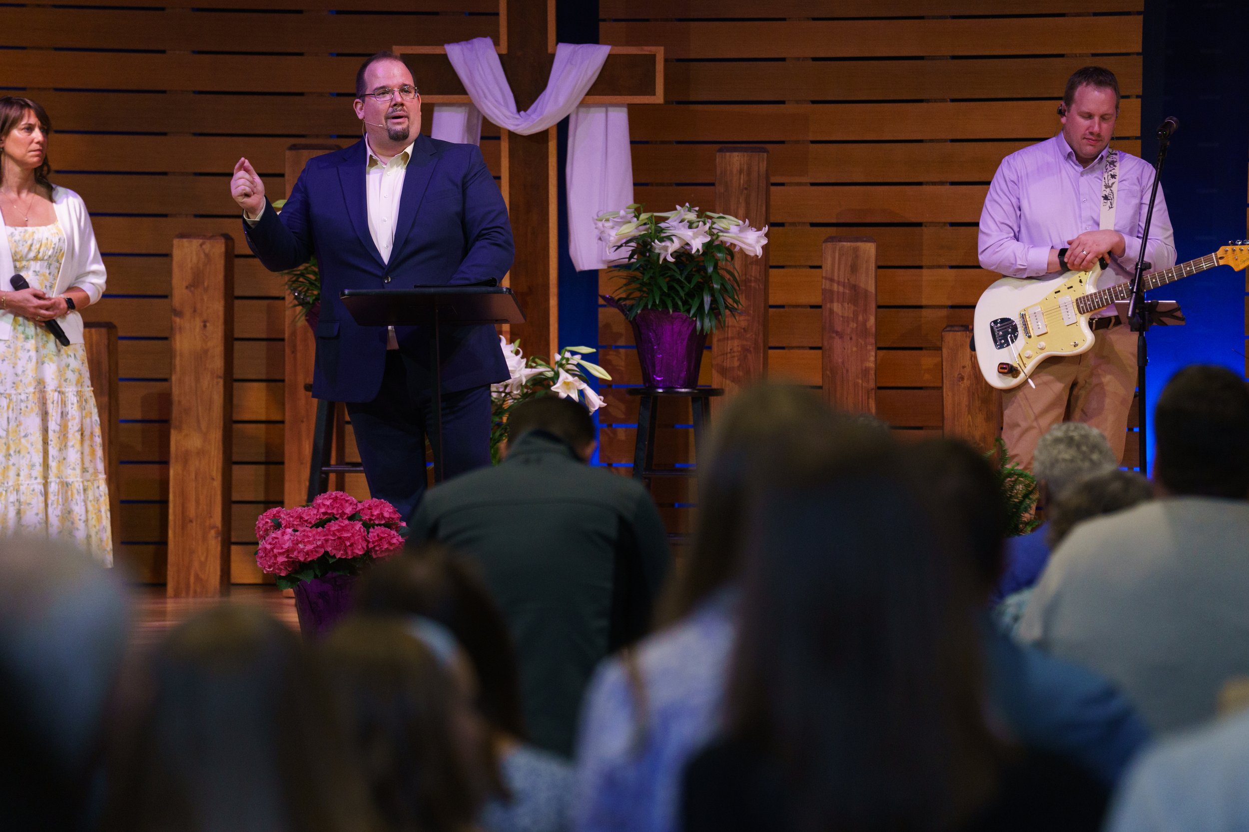 People gathered at a church service. One man in a blue suit is speaking at the pulpit, a woman holds a microphone, and another man on guitar stands nearby. The church interior features wooden panels and decorative flowers at Easter Service.