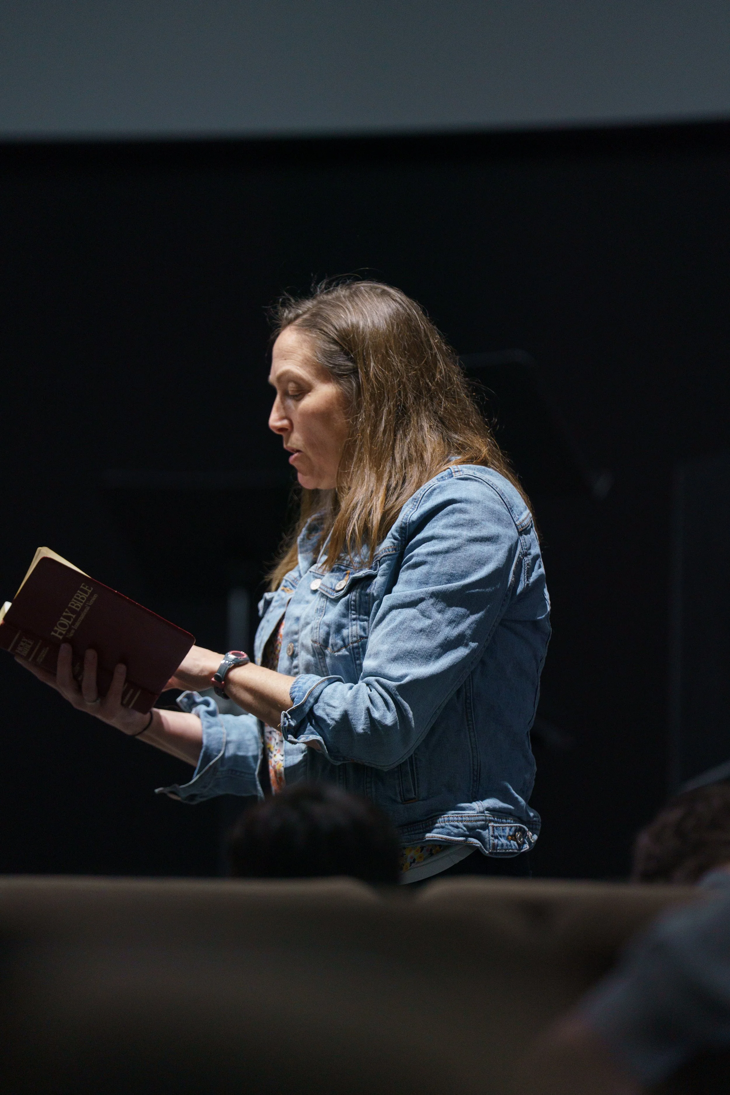A woman with long brown hair wearing a denim jacket is reading a book titled 'Holy Bible' in a children's church class at Victory Life Church in Stow, OH, near Tallmadge, OH.