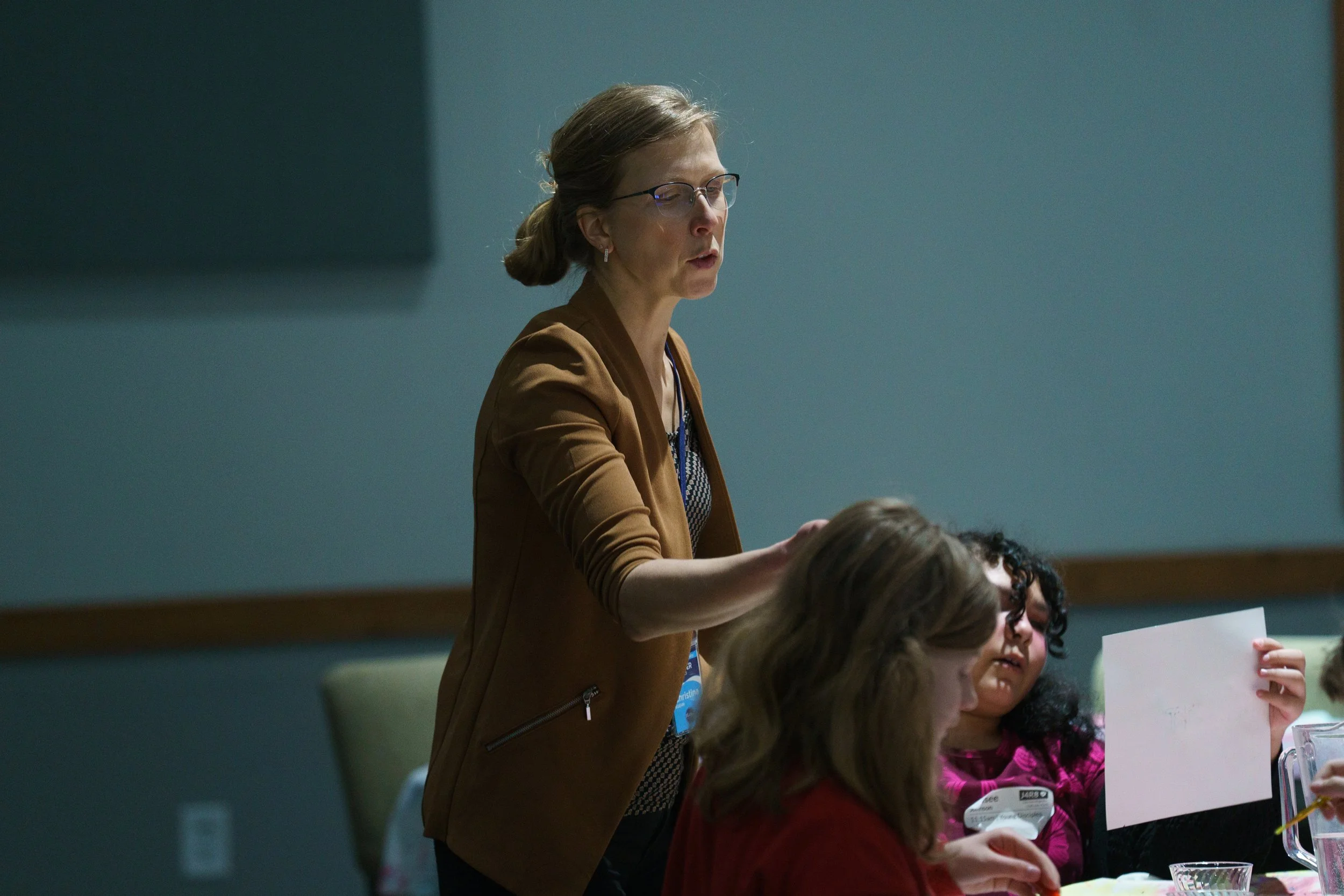 A woman teaching a children's church class, wearing glasses and a brown jacket standing at a table with children, speaking and gesturing as she teaches at Victory Life Church near Cuyahoga Falls, OH.